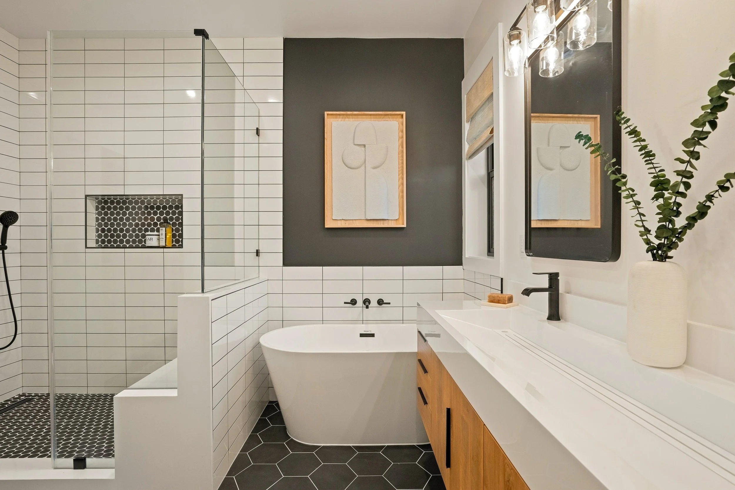 Modern bathroom with white subway tiles, a black and white hex tile shower floor, a framed abstract art piece on a dark wall, a white bathtub, wooden vanity with white countertop, black faucet, and a greenery plant next to a mirror with glass light f
