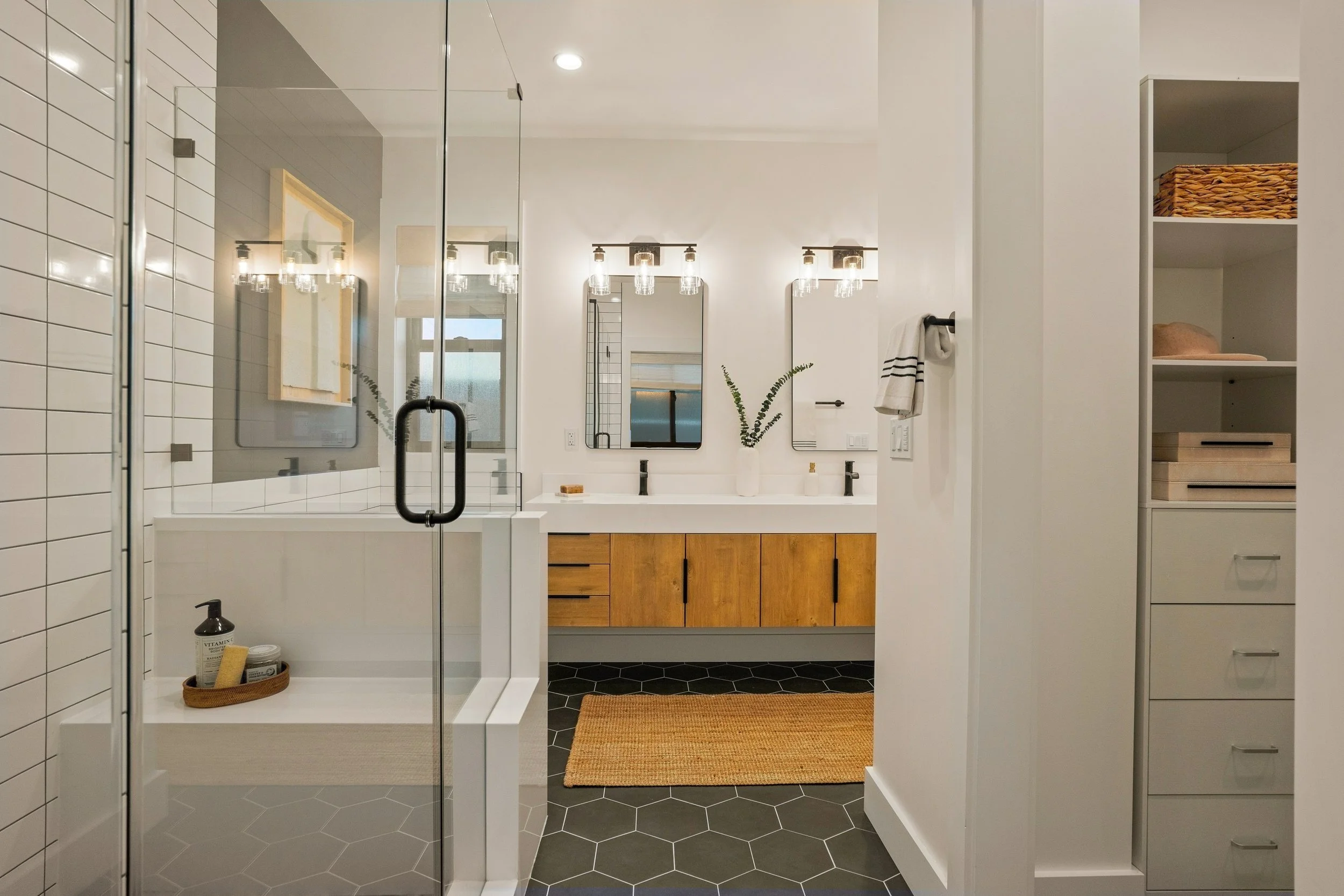Modern bathroom with white subway tile walls, a glass shower door, black hexagonal floor tiles, a double vanity with wood cabinets, a mirror with lighting, and open storage shelves with woven baskets and hats.