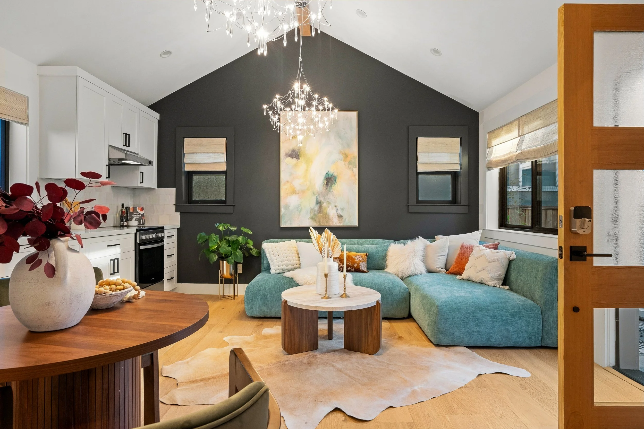 Living room with a teal sectional sofa, a large abstract painting, and a black accent wall. Chandelier hanging from the ceiling. Multiple windows with beige blinds. Wooden furniture and plants.