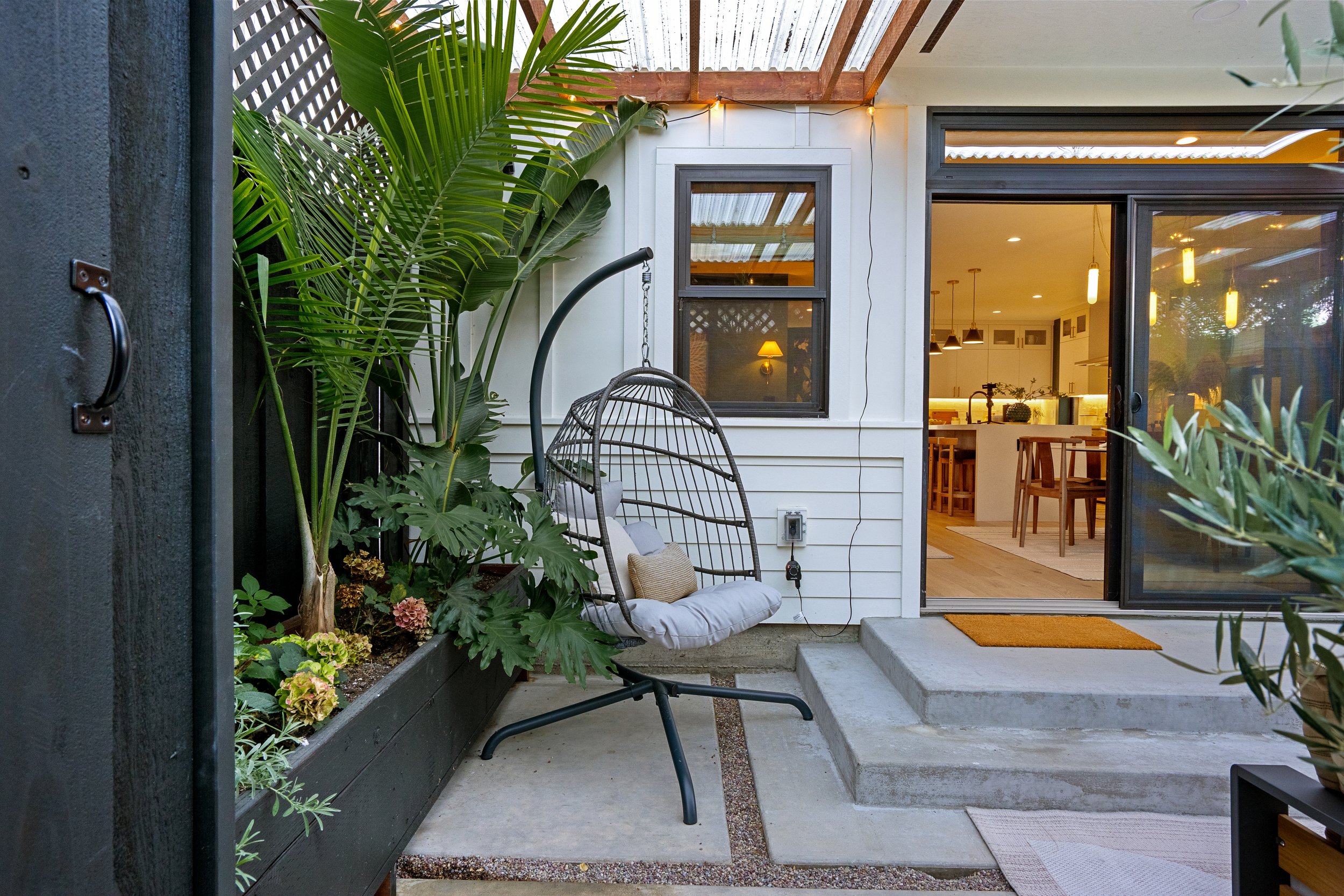 A backyard patio area with large green leafy plants, a hanging chair with beige cushions, and a sliding glass door leading to a lit interior dining space.