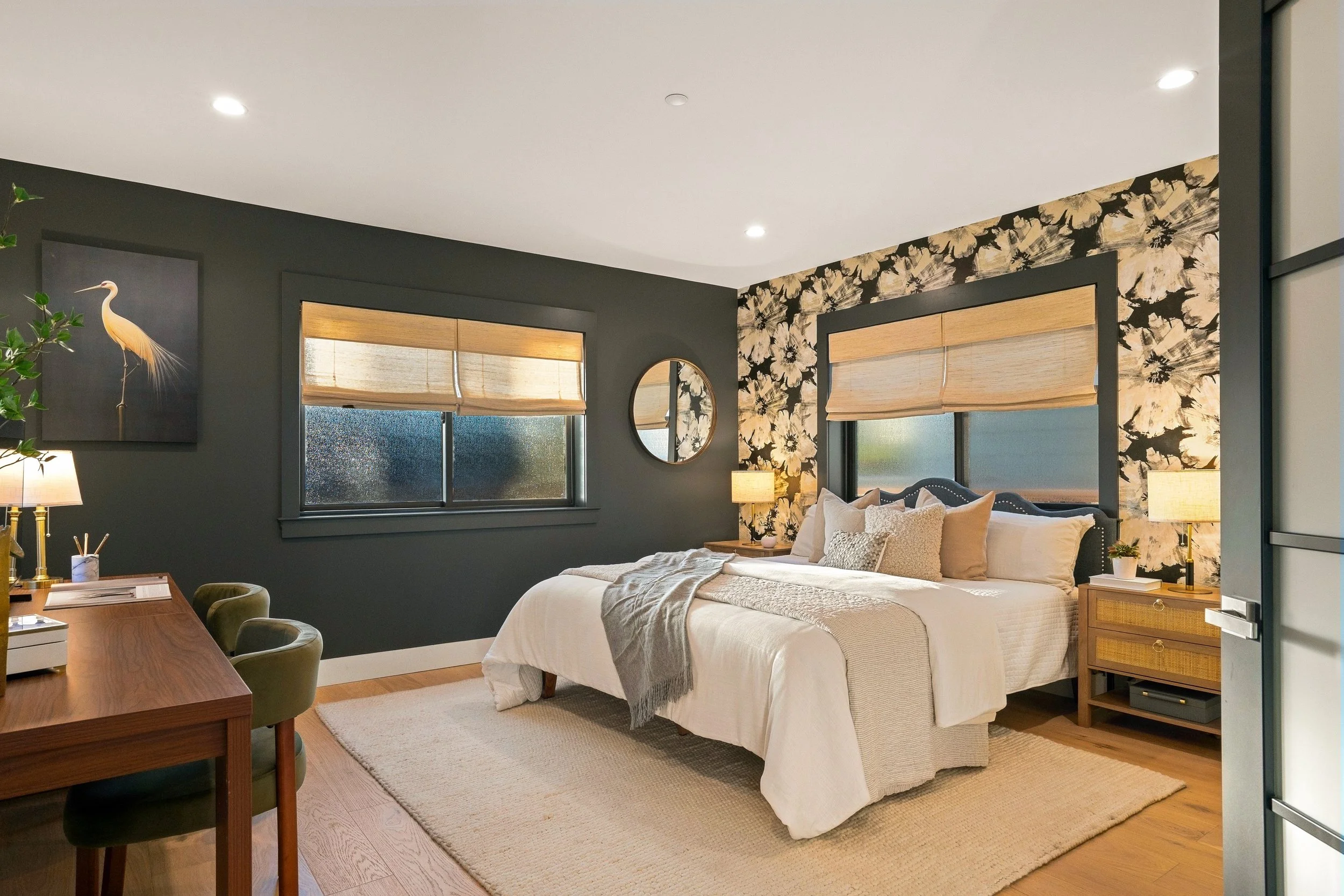 A bedroom with a bed, pillows, and blankets, dark gray walls with floral wallpaper, two windows with beige blinds, a round mirror, nightstands, lamps, and a desk with chairs, decorated with plant and artwork.