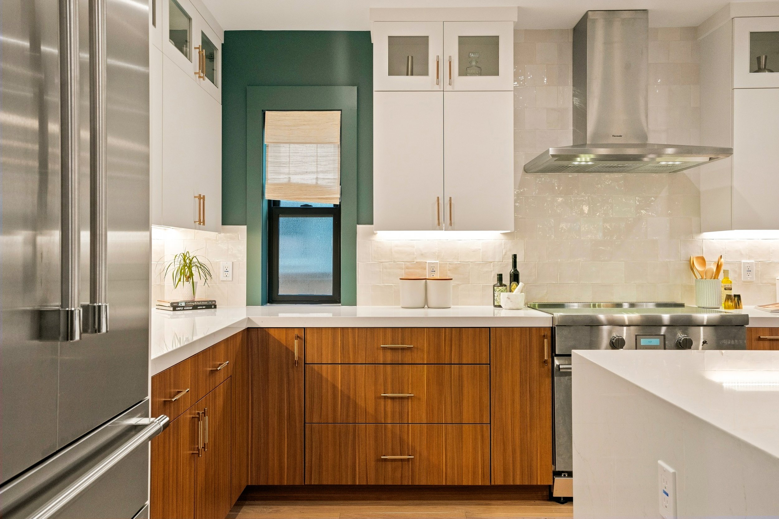 Modern kitchen with white upper cabinets, wooden lower cabinets, stainless steel appliances, and a window with a bamboo shade.
