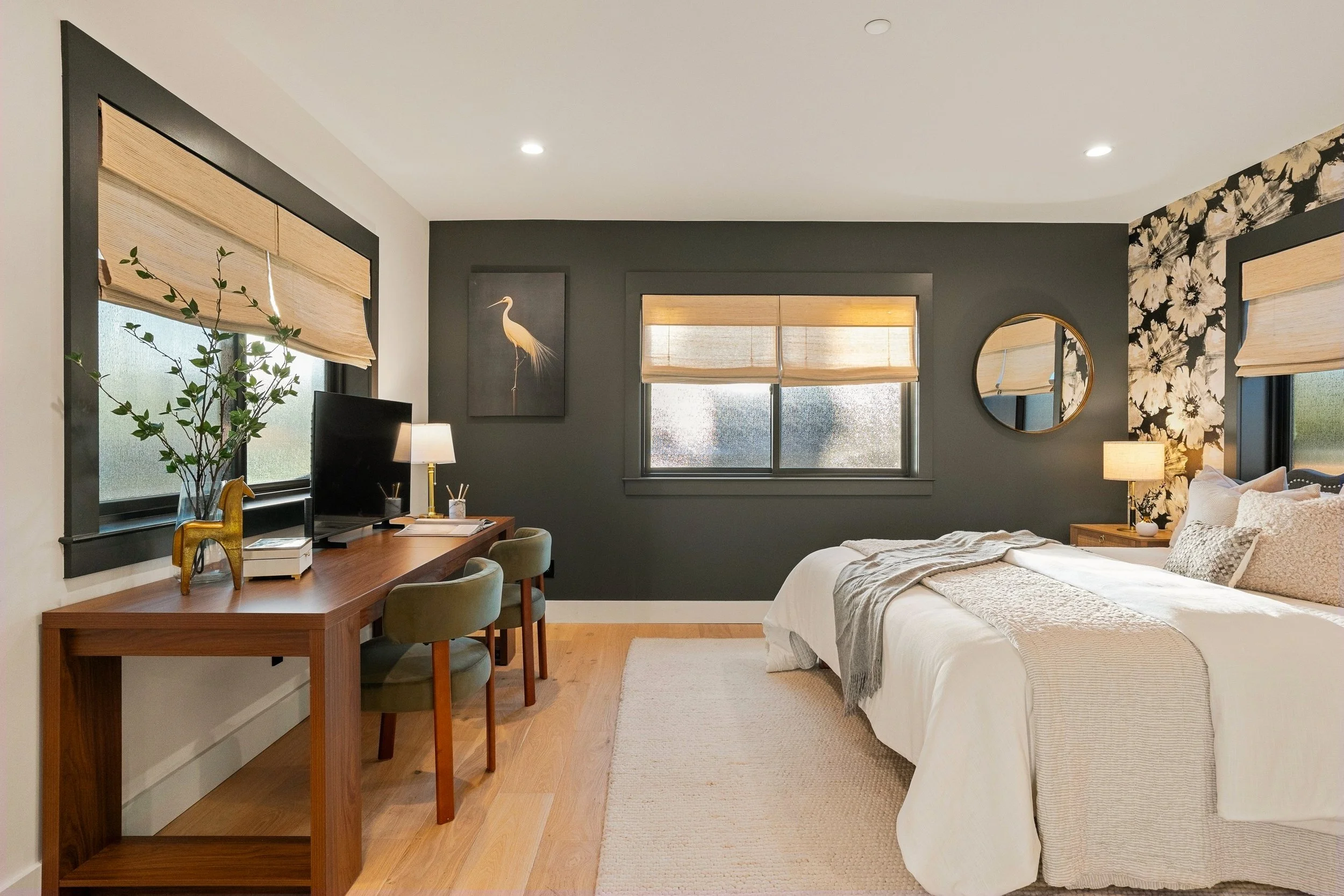 A bedroom with dark gray walls, three windows with bamboo blinds, a white bed with patterned pillows, a wooden nightstand with a lamp, a round mirror, floral wallpaper on one wall, a wooden desk with a computer, books, a small lamp, and a plant, and 