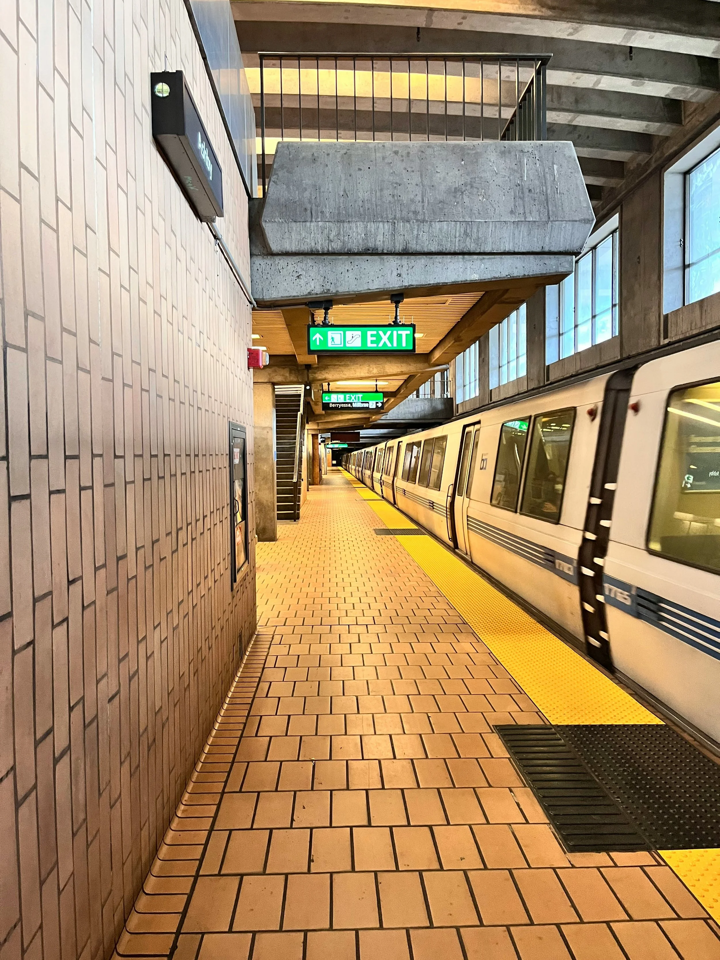 An underground subway station platform with a train on the right side, illuminated exit signs hanging from the ceiling, brick flooring, and large windows on the right. There is a staircase in the background and a beige tiled wall on the left.