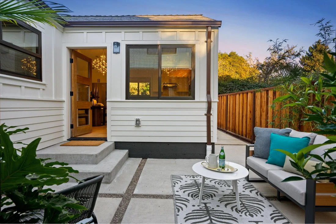 Small backyard patio with outdoor seating, a white patterned rug, a black and white loveseat with blue and gray pillows, a small round table with drinks, and a wooden fence, adjacent to a house with white siding and a glass door.
