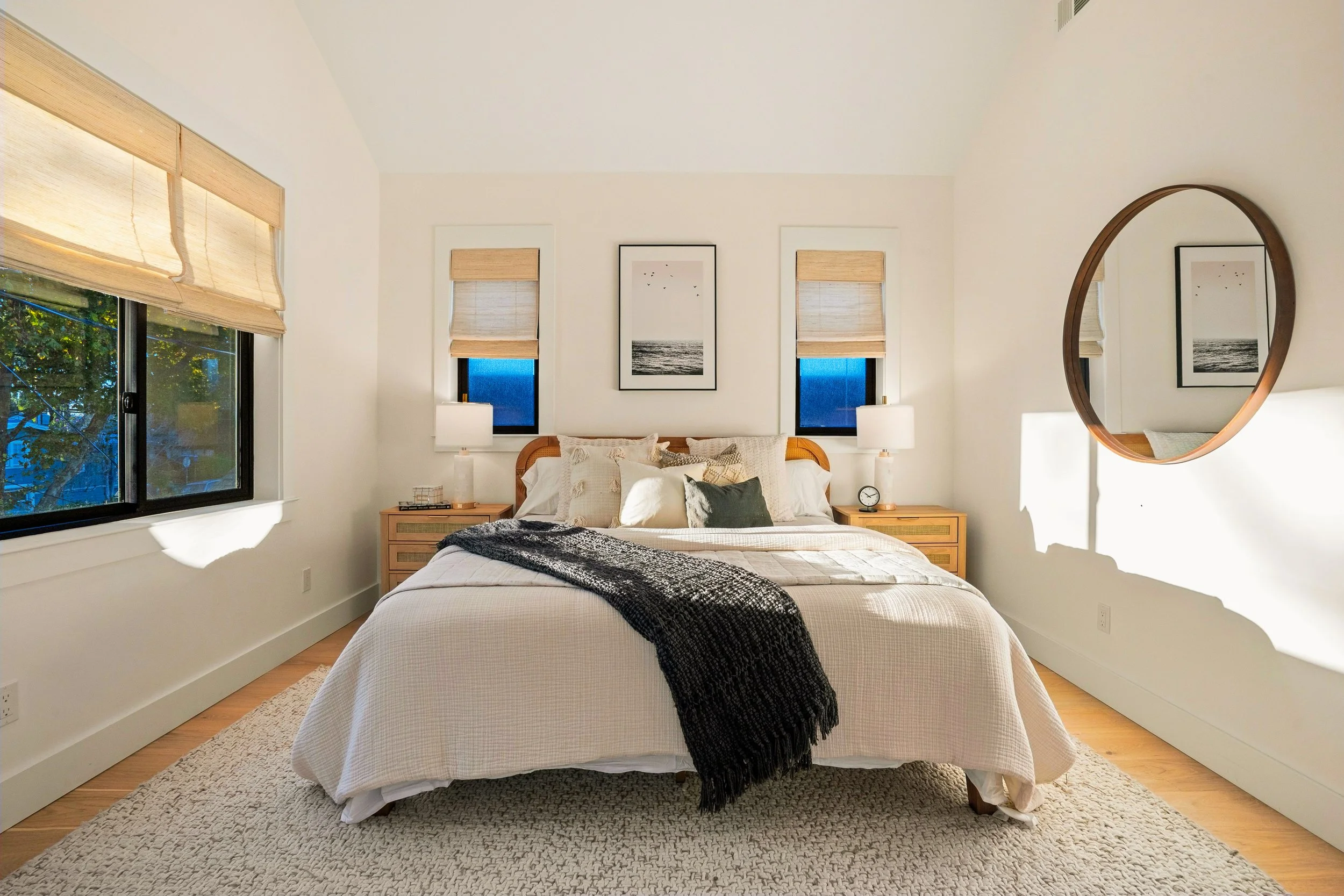 A cozy bedroom with a window, bed, nightstands, lamps, framed artwork, a large round mirror, and a textured rug.