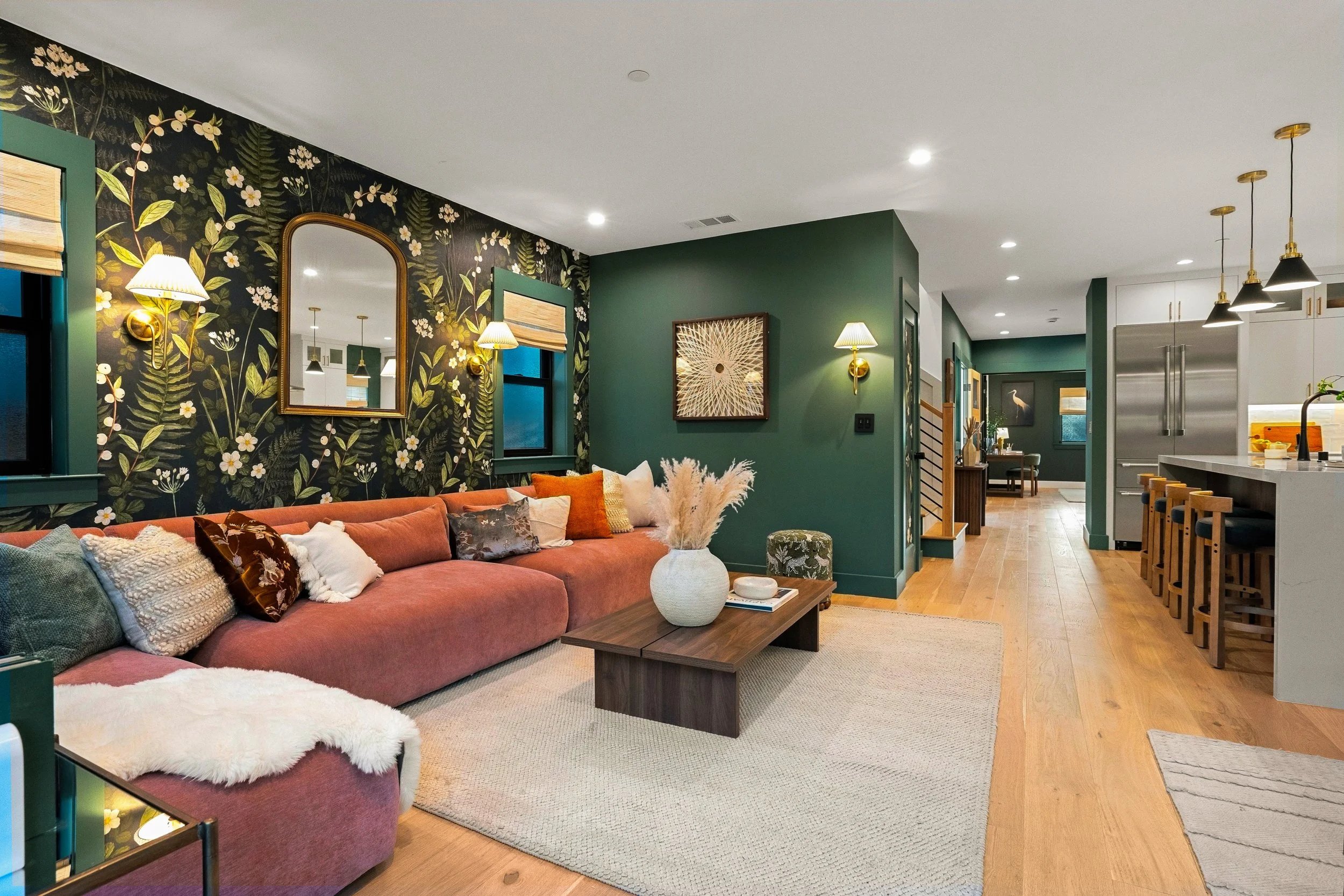 A living room with a pink sofa with various pillows, a coffee table with a white vase, a wall decorated with floral wallpaper, gold and white sconces, and a framed piece of art on a green wall. Part of a kitchen with white cabinets, a stainless steel