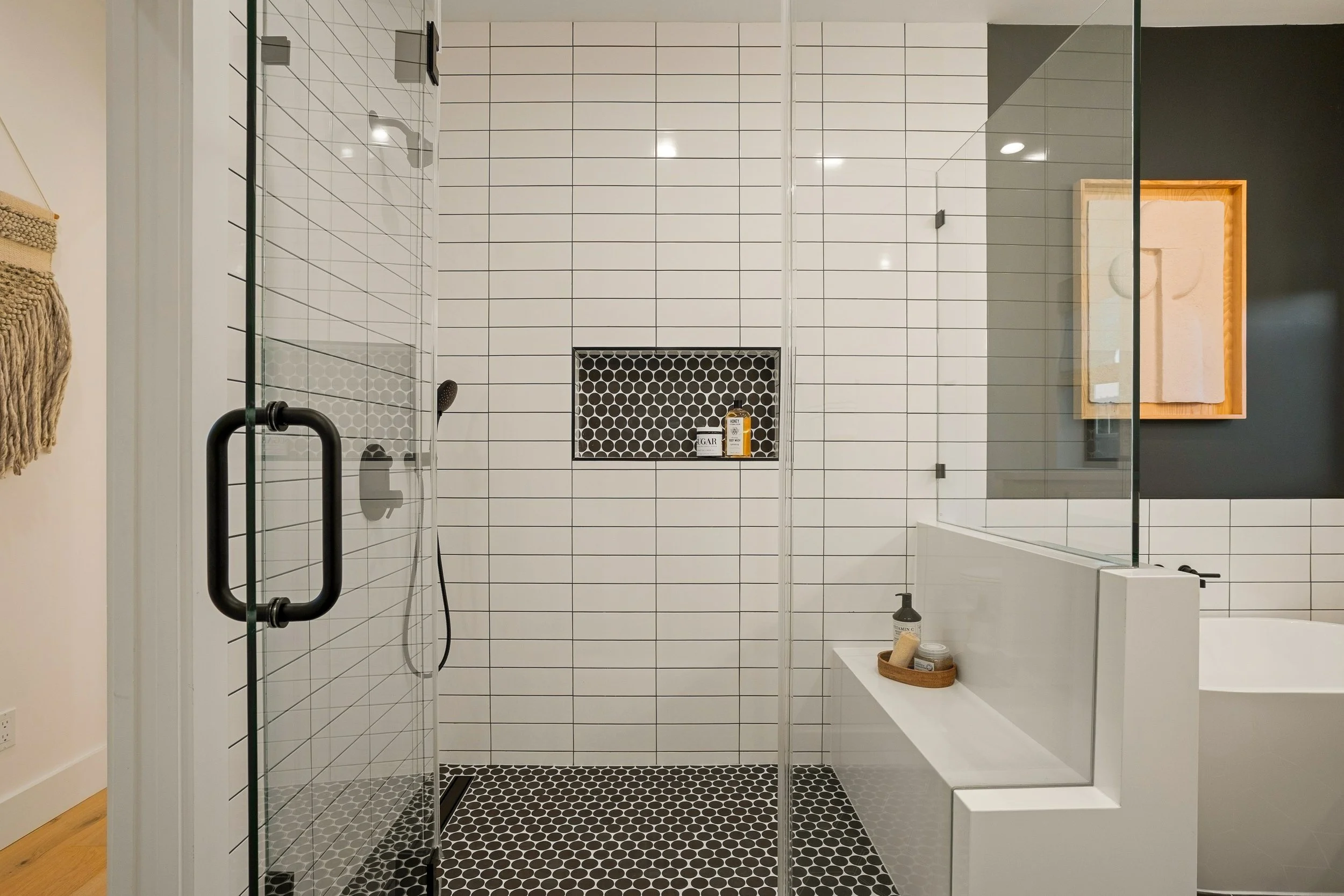 A modern walk-in shower with black and white hexagonal floor tiles, white subway-tile walls, a built-in niche with black tiles holding bath products, a glass shower door with black hinges, and a white bench. The shower has a black showerhead and a ha