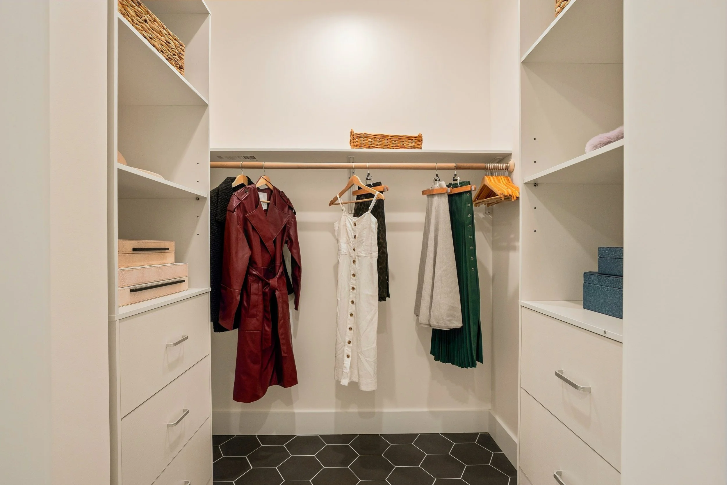 Empty walk-in closet with white shelving and hanging clothes, including a red jacket and a white dress, and a basket on top shelf.