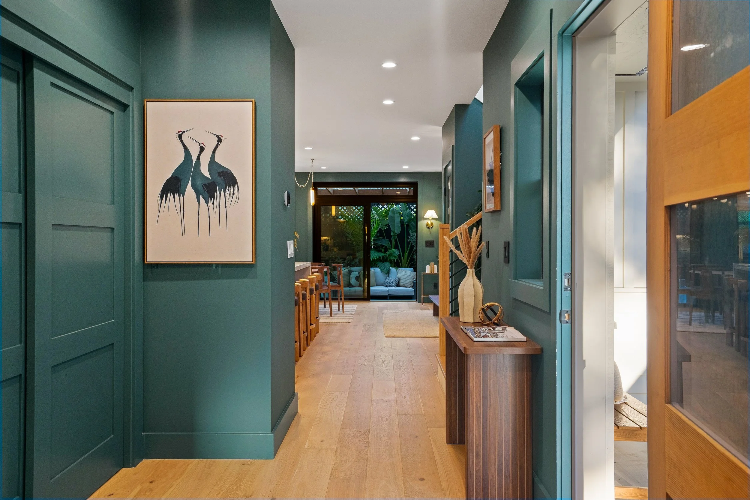 Interior view of a modern home with green and wooden walls, artwork of three cranes hanging, a staircase, and a living room with large windows and lush greenery outside.