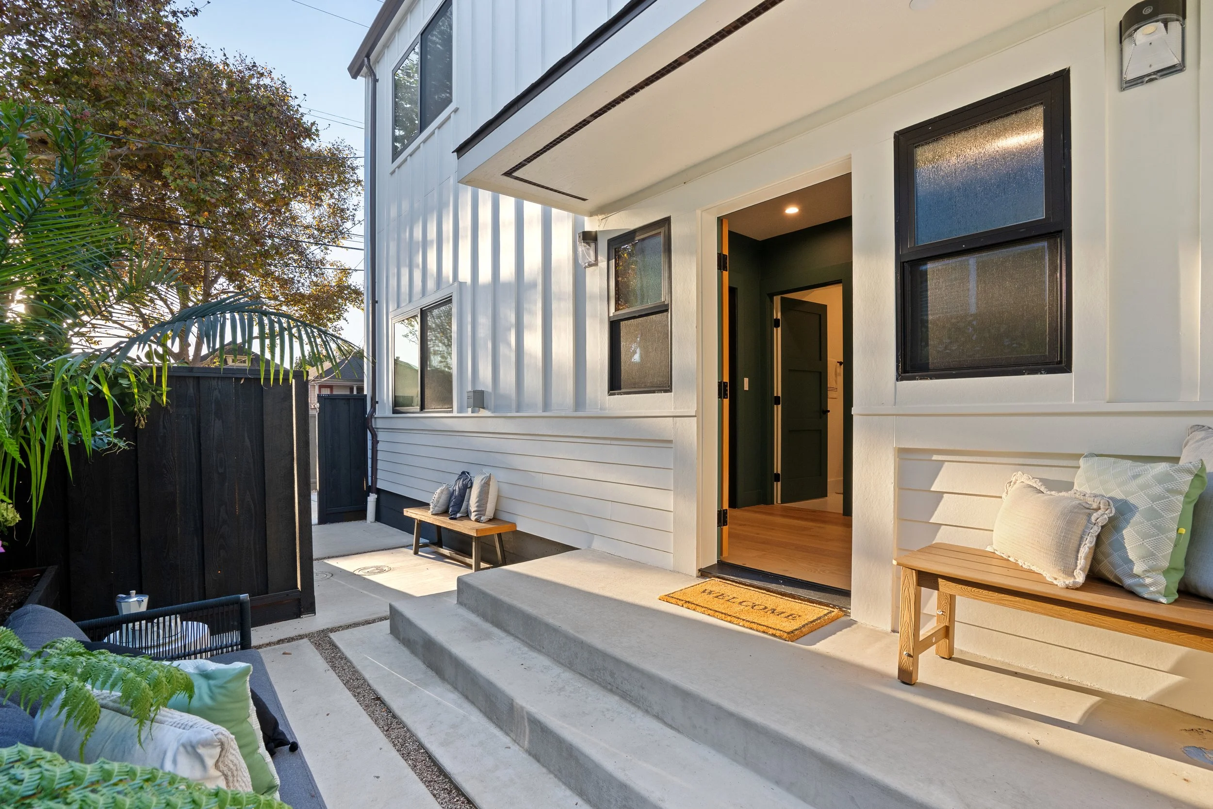 A modern house with an open door leading to a living room, a welcomed mat at the entrance, outdoor seating with cushions, and a small wooden bench on a concrete porch surrounded by lush plants and trees.