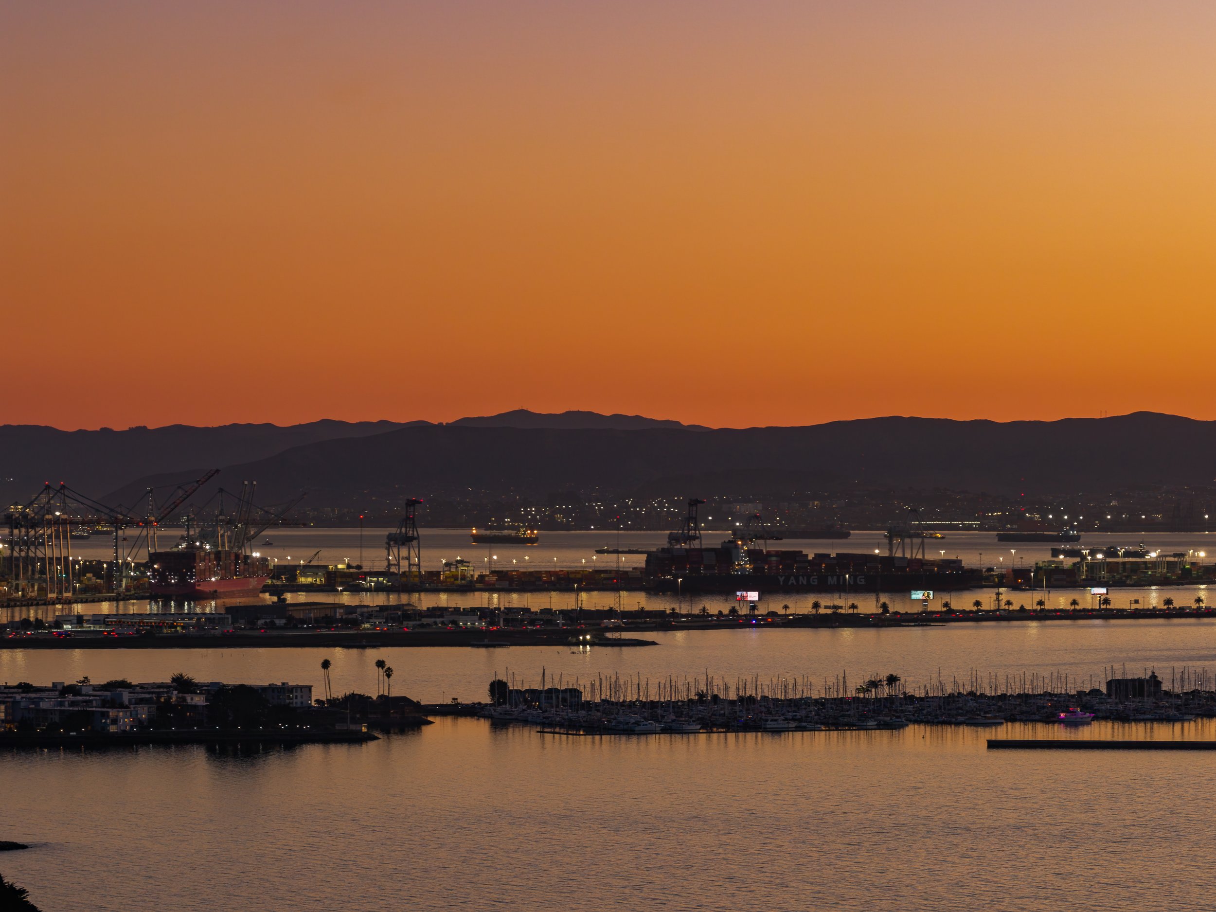 A scenic sunset view over a harbor with sailboats, industrial cranes, and distant hills, reflecting the warm colors of the sky.