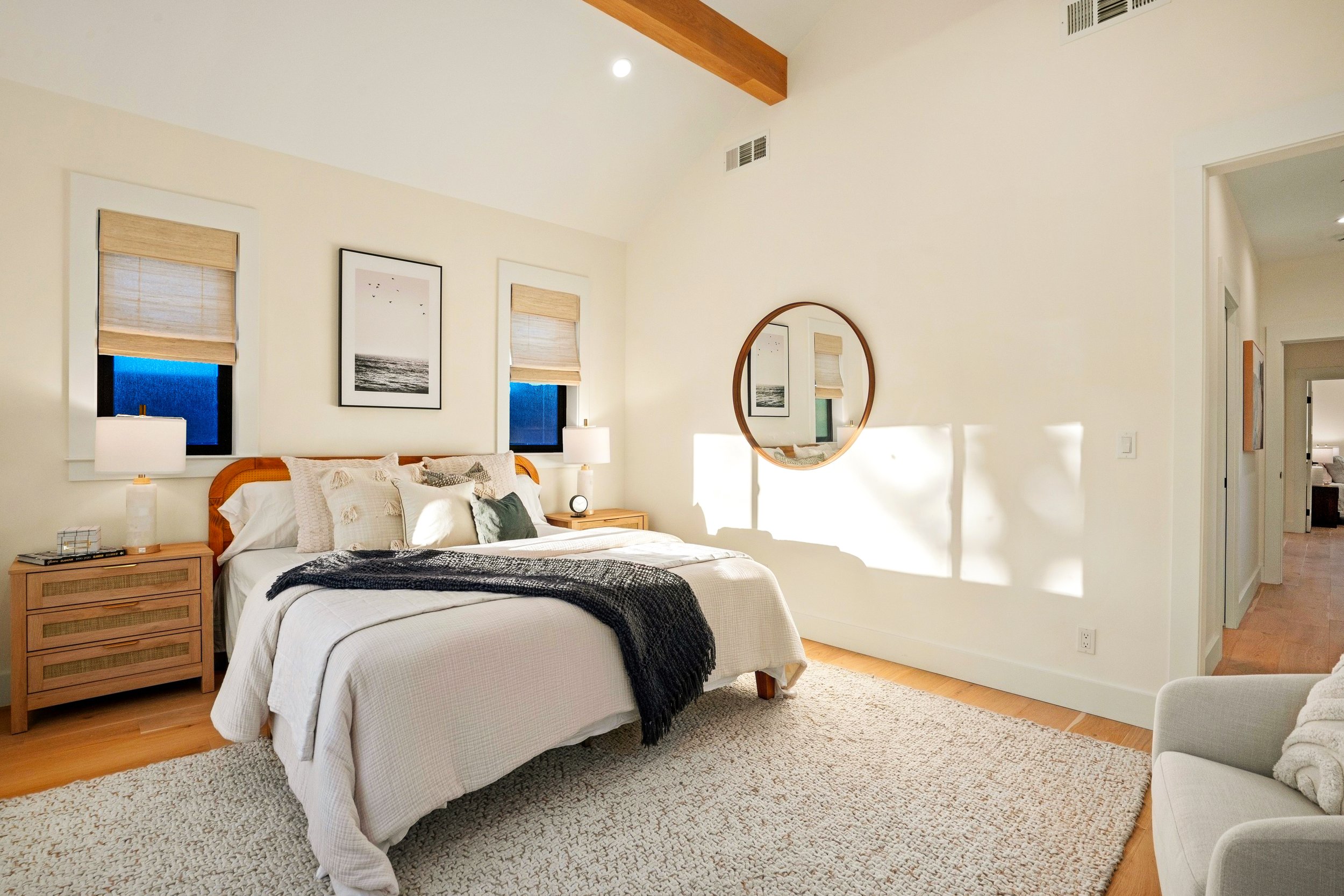 A bedroom with a bed, two nightstands with lamps, framed artwork, a round mirror, beige walls, hardwood floors, and natural light coming through two windows.