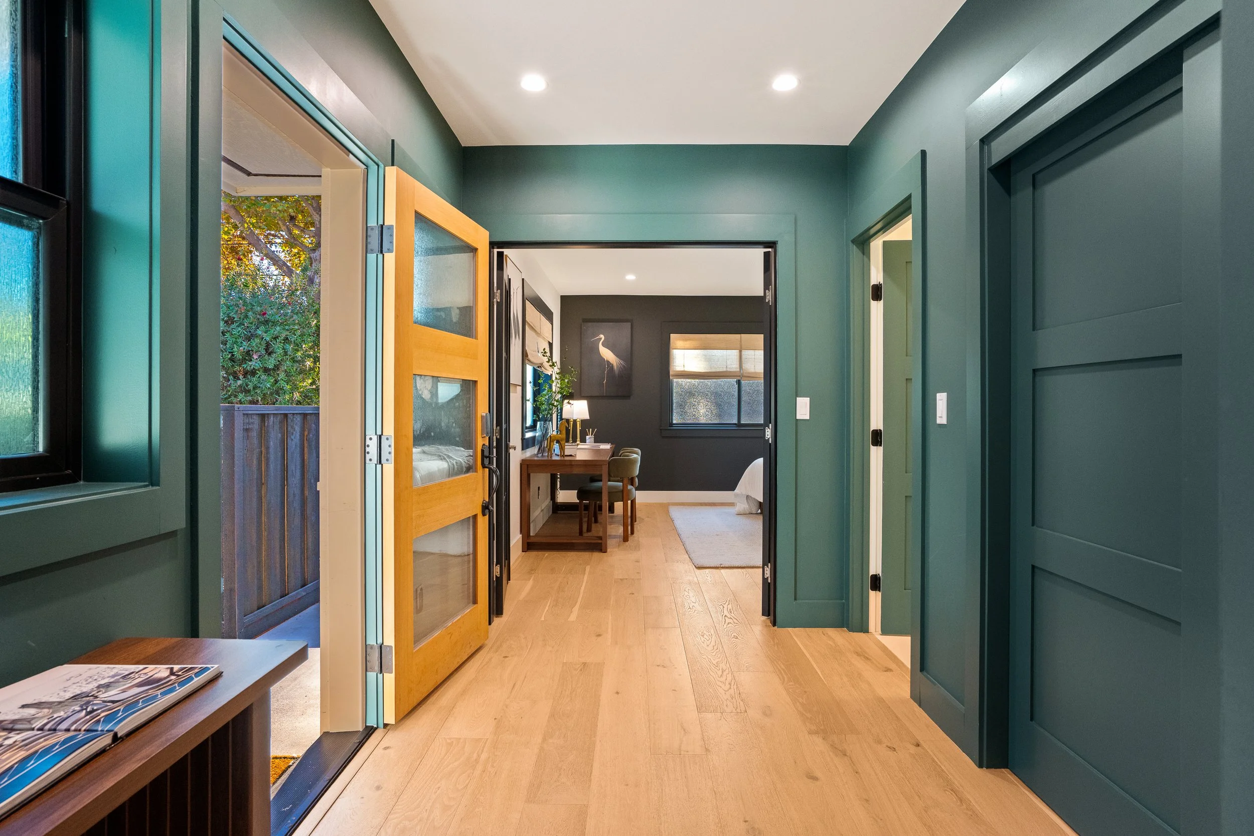 Interior view of a house entryway with green walls, wooden flooring, and an open door leading to a living room with a desk, chairs, and artwork.
