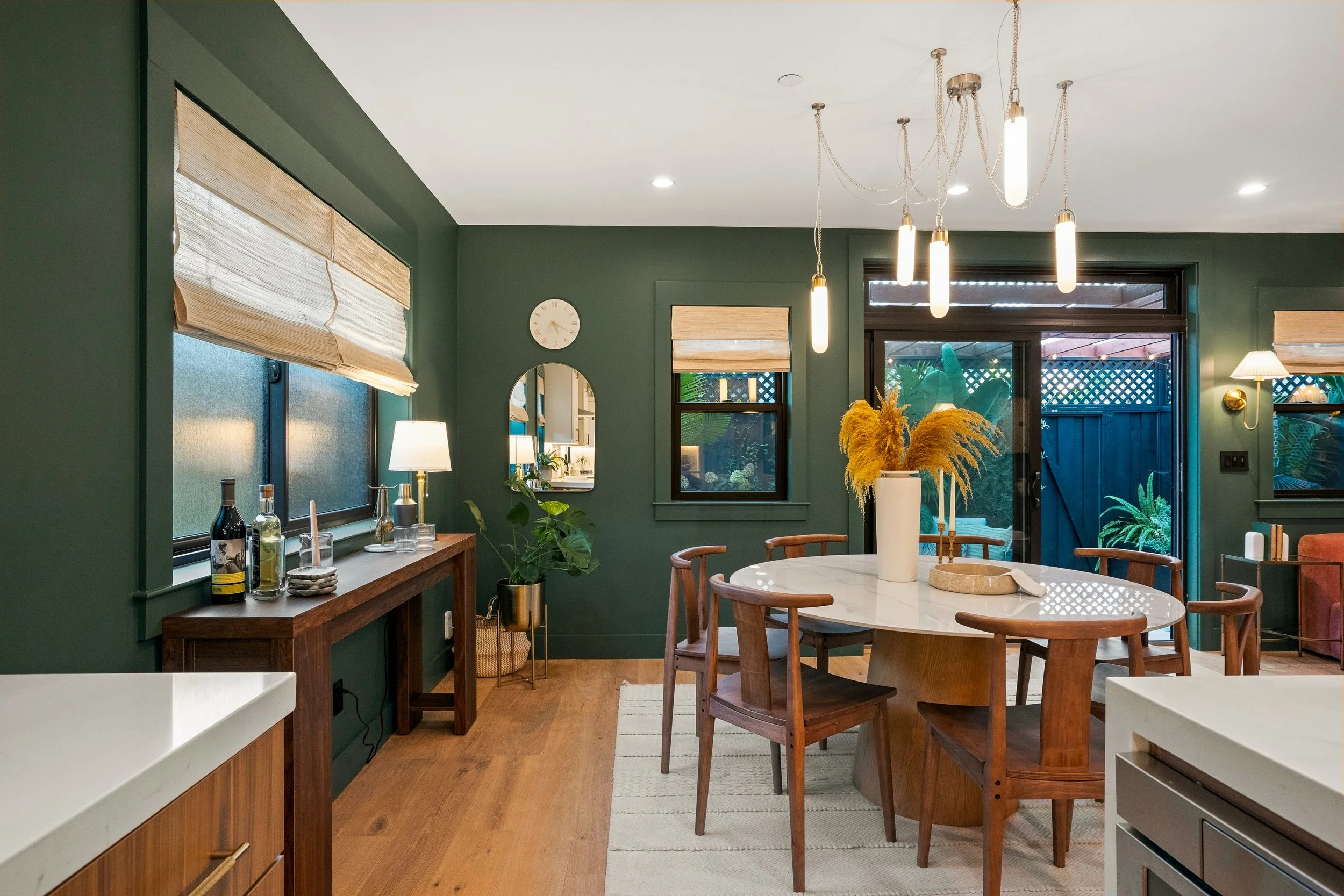 A modern green kitchen and dining area with a round white marble table, wooden chairs, and pendant lights hanging from the ceiling. There are windows with beige blinds, and a wooden sideboard with bottles and glasses. The room has wooden flooring, an