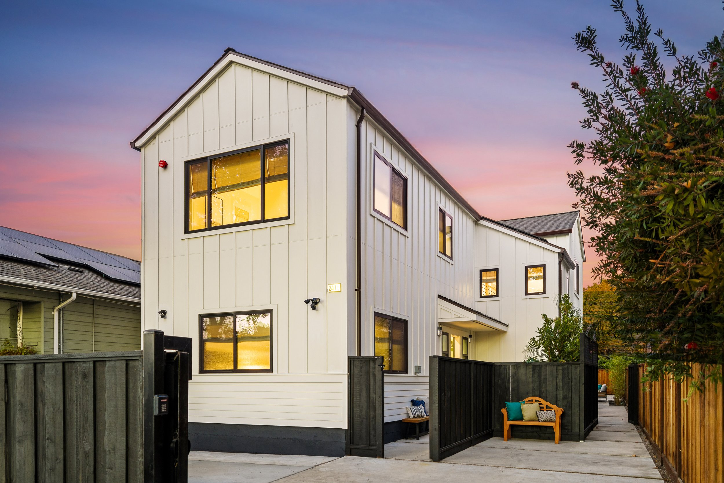 Modern white multi-story house with large windows at sunset, surrounded by a black fenced yard, with a small porch area with seating, and a tree on the right side of the image.