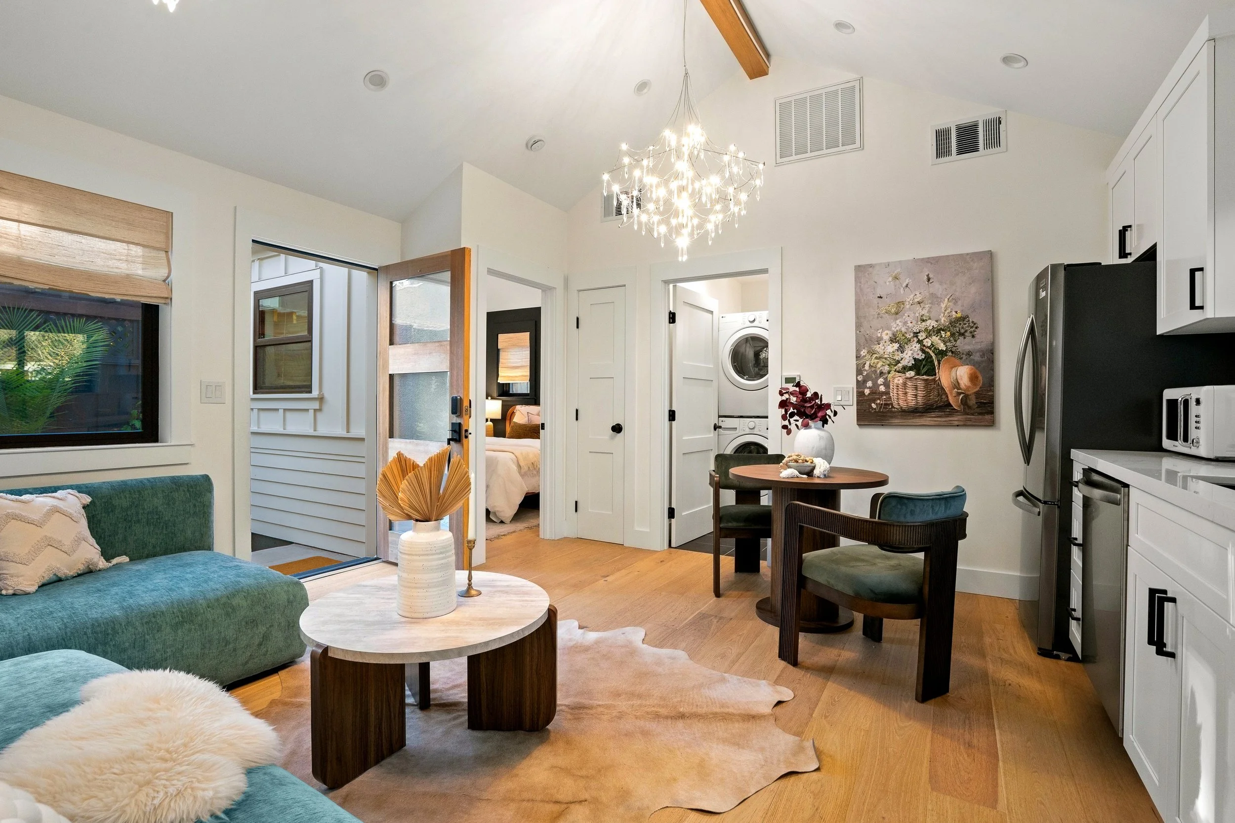 An open-concept living space with a green sofa, coffee table, and a fluffy sheepskin rug in the foreground. The room has wooden flooring, a wall with windows, and a chandelier hanging from the ceiling. There is a view into a bedroom with a bed and a 