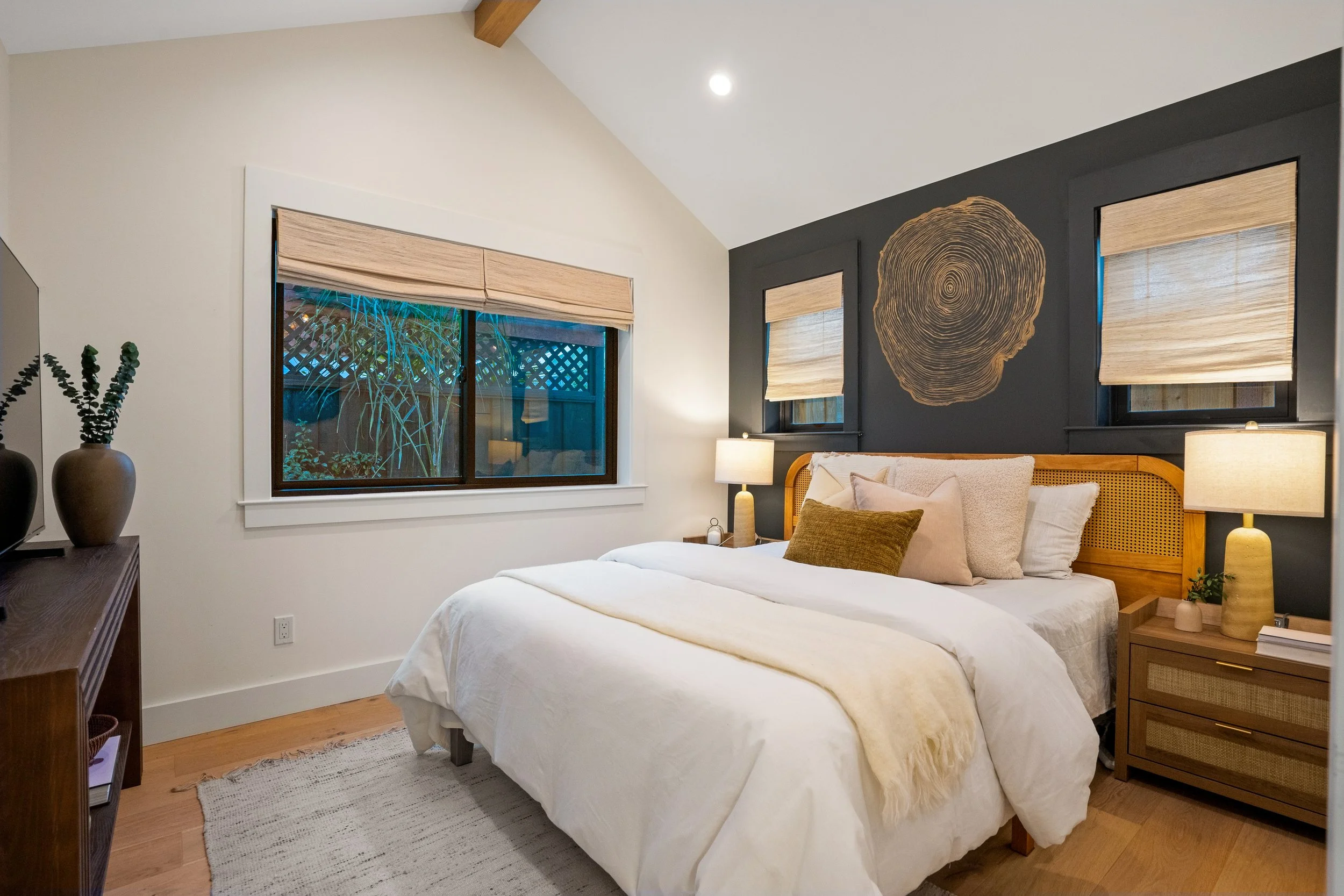 A bedroom with a bed, nightstands, lamps, and two windows with blinds. One window is on a dark wall with a wooden wall art piece resembling a tree cross-section. The room has a wooden floor, a dresser with vases, and a small plant.