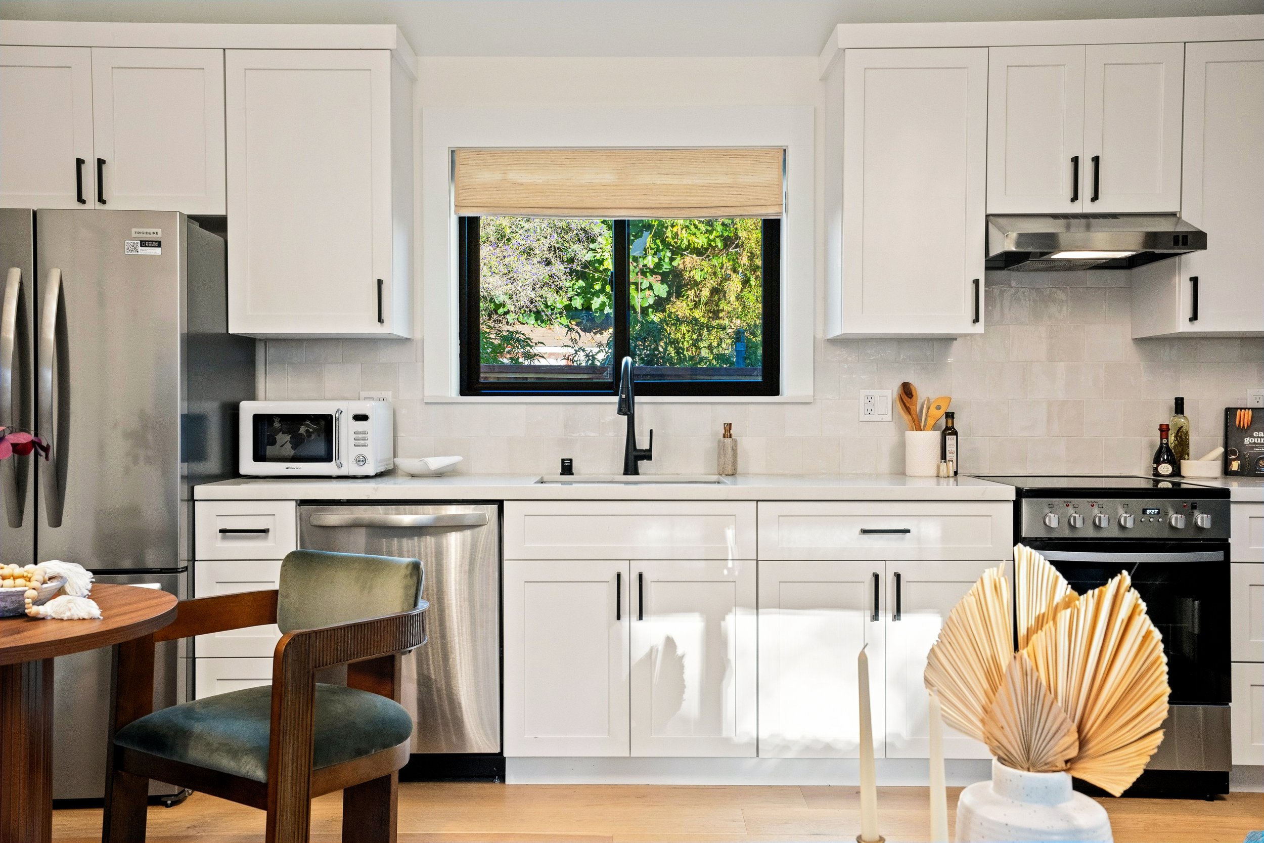 Modern kitchen with white cabinets, a window showing green trees outside, black handles on cabinets, and a stainless steel refrigerator. A wooden table and chairs are in the foreground, with decorative items on the table and a offset paper sculpture 