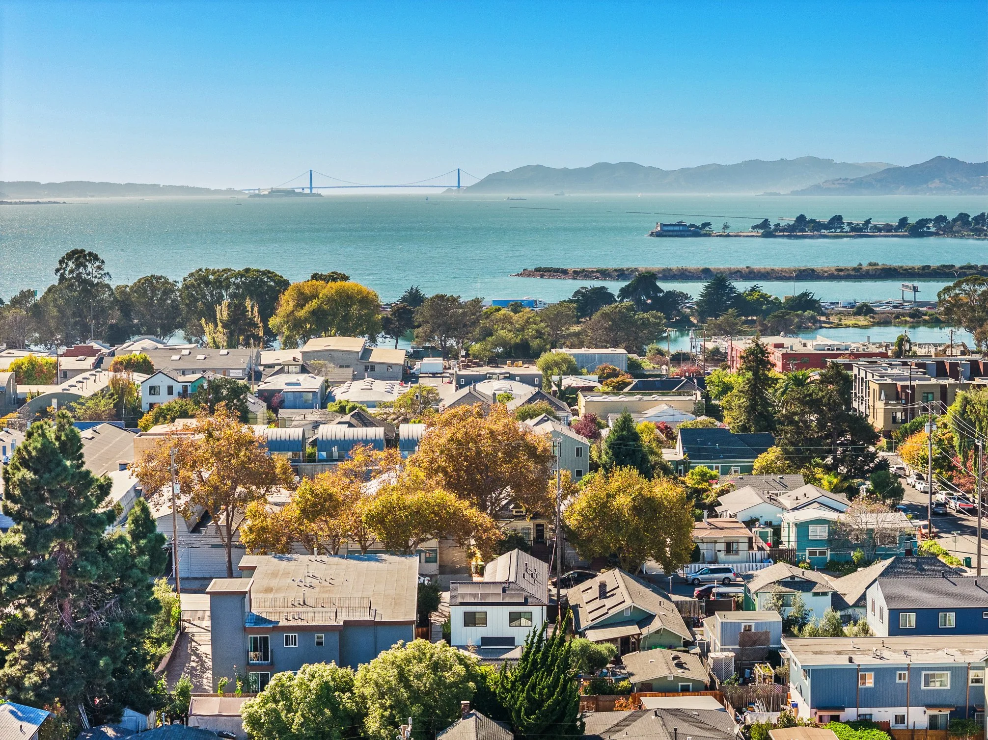 A picturesque view of a residential neighborhood with houses and trees in the foreground, a body of water with boats in the middle ground, and a bridge with hills in the background under a clear blue sky.