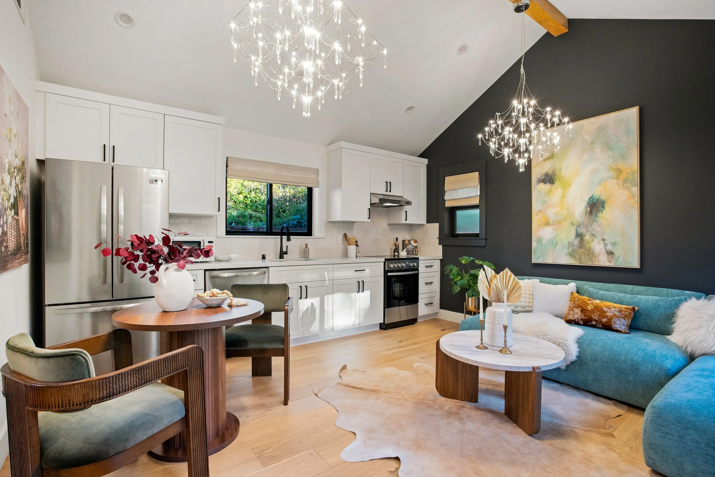 Open-concept living room and kitchen with modern decor, white cabinets, stainless steel appliances, black window frame, dark accent wall with large abstract painting, chandelier lights, a teal sofa with pillows, wooden coffee tables, and a small roun