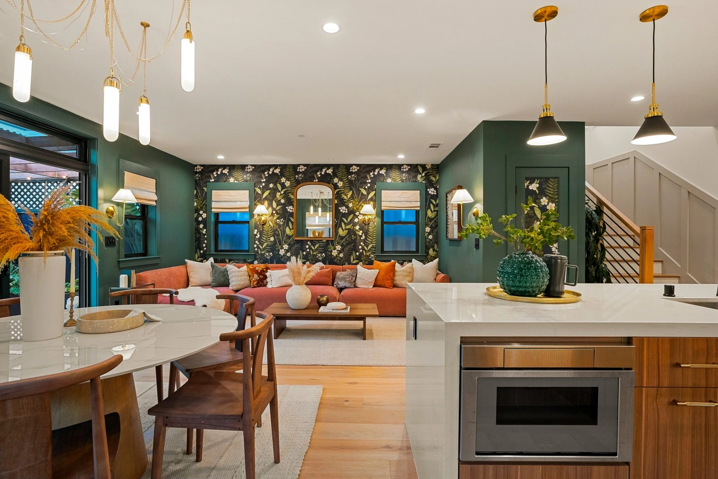 Modern open-concept living space featuring a kitchen with a marble island, a coral-colored sectional sofa, decorative vases, framed art, and a dark floral wallpaper accent wall with three windows and illuminated by stylish pendant and wall sconces.