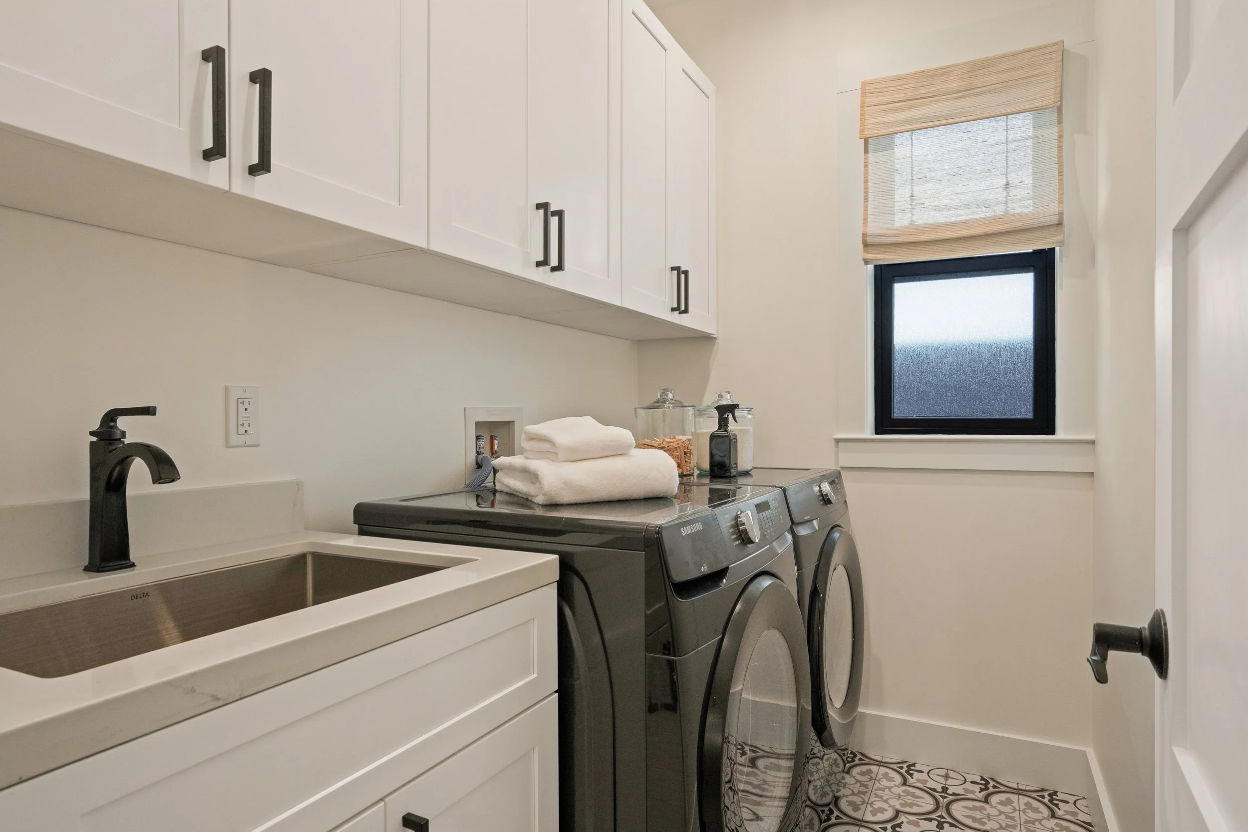 Laundry room with white cabinets, a black sink faucet, a washing machine and dryer, a small window with a bamboo shade, and a tiled floor.