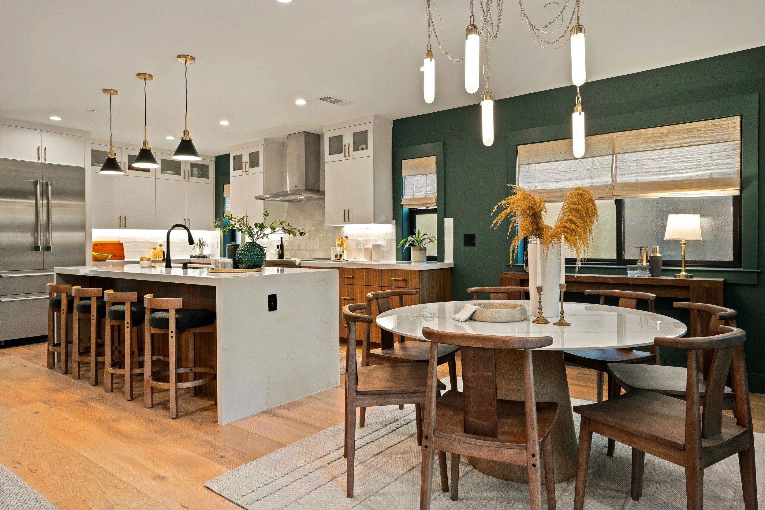 Modern kitchen with white cabinets, stainless steel appliances, a large island, and a dining area with a round table and wooden chairs. Decor includes plants, a vase with dried flowers, and a table lamp. Pendant lighting hangs over the island and din