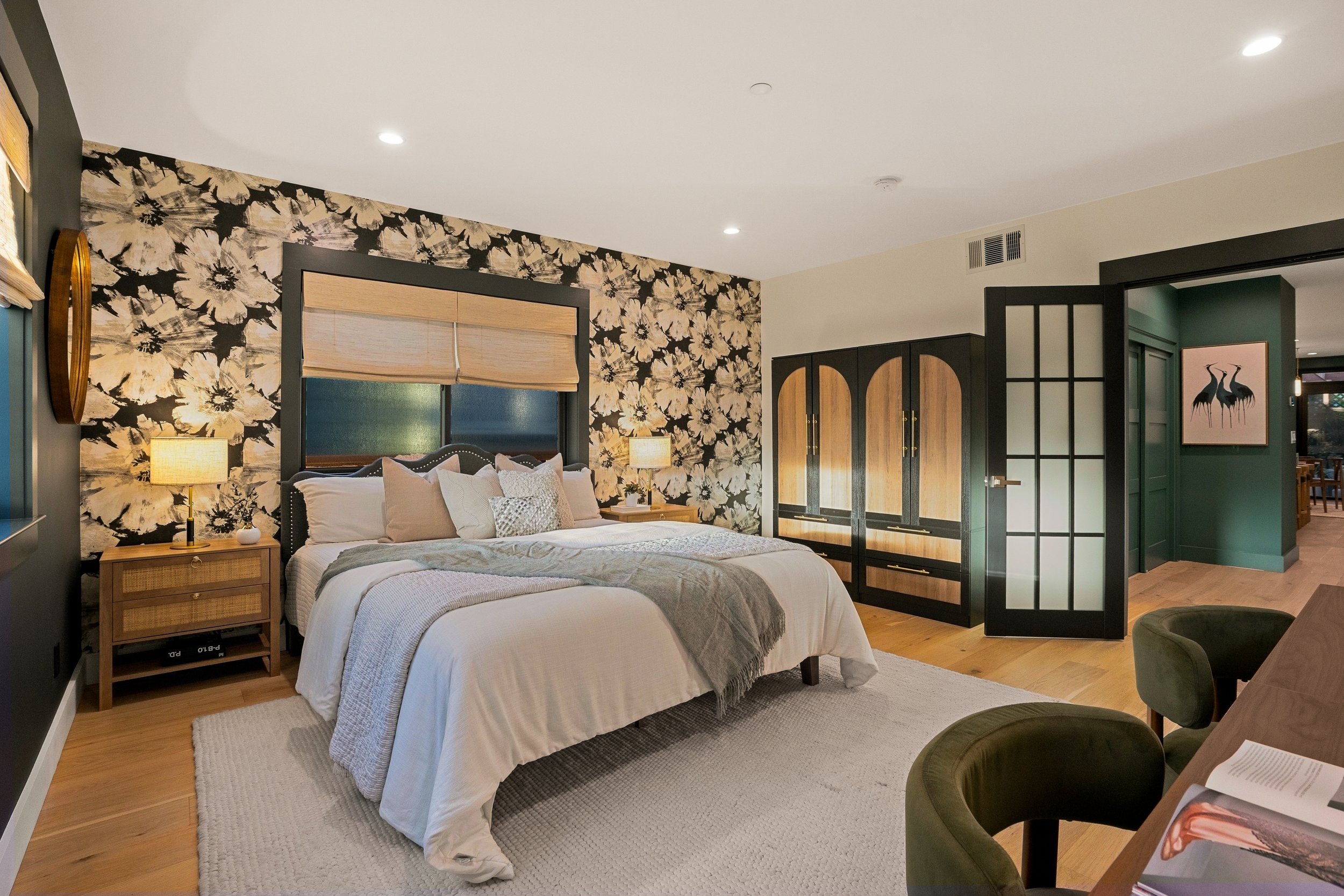 A bedroom with floral wallpaper behind a bed with beige and white linens. There are two bedside tables with lamps, a closet with wood and black doors, and a green wall with a framed artwork of two herons. The room has hardwood floors and recessed cei