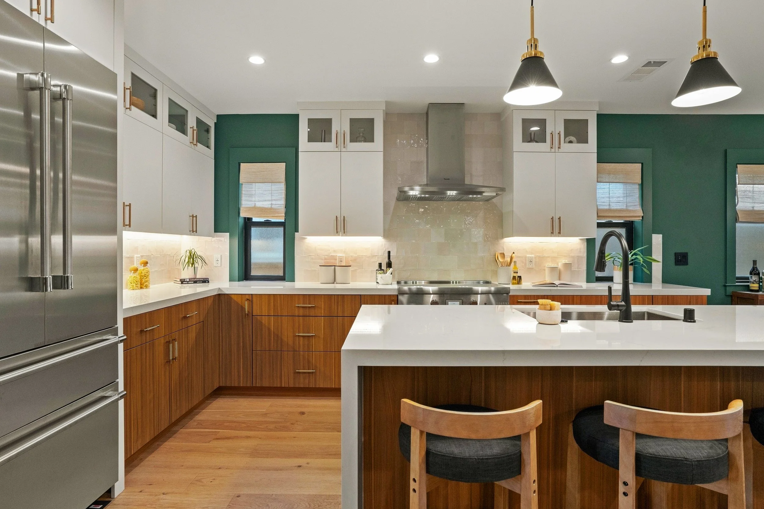 Modern kitchen with white and wooden cabinetry, stainless steel appliances, a large island with a white countertop, black sink, and wooden chairs, green accent walls, and pendant lighting.