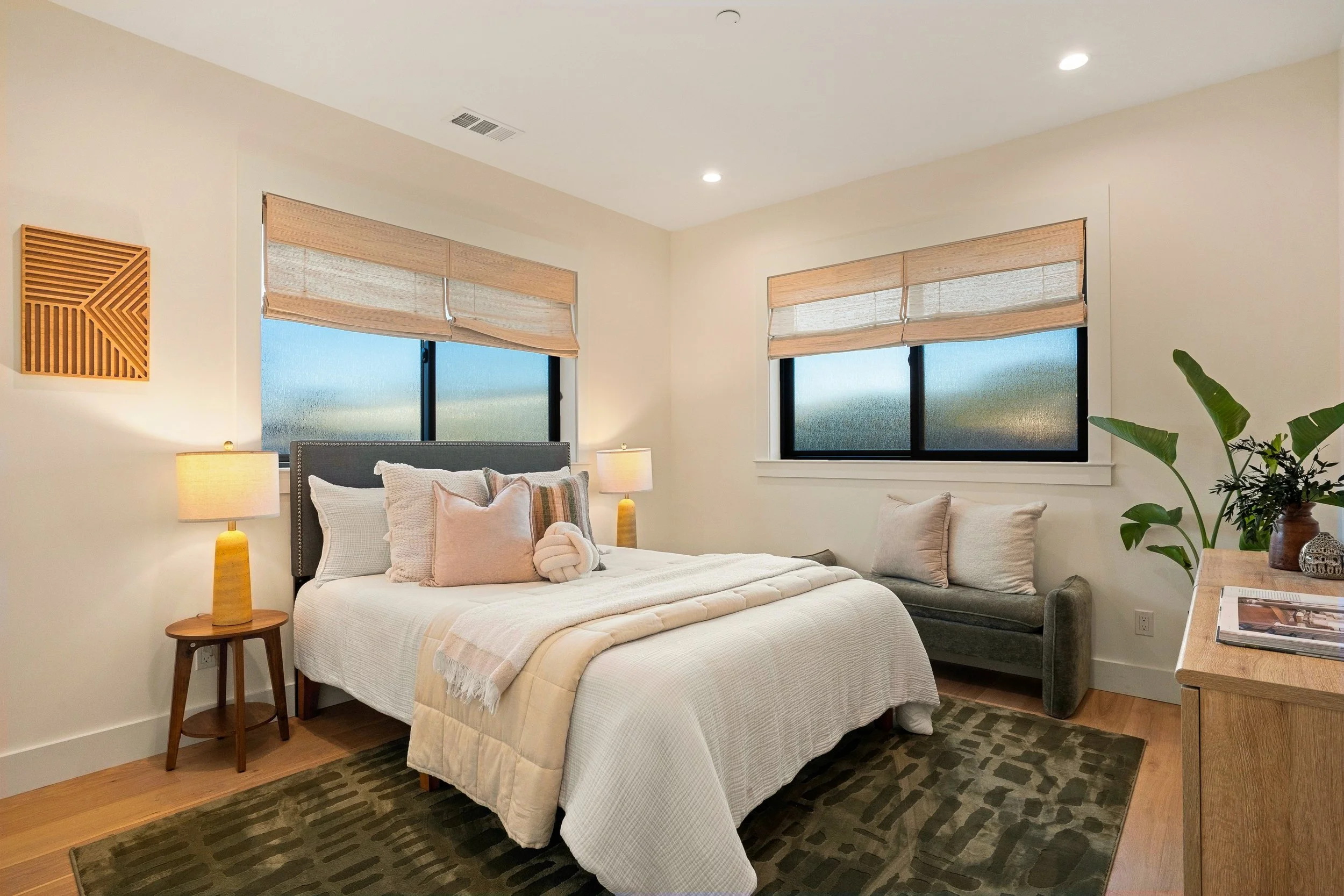 A bedroom with a bed, multiple pillows, two nightstands with lamps, and large windows with beige blinds. There is a green plant and a wooden side table with books.