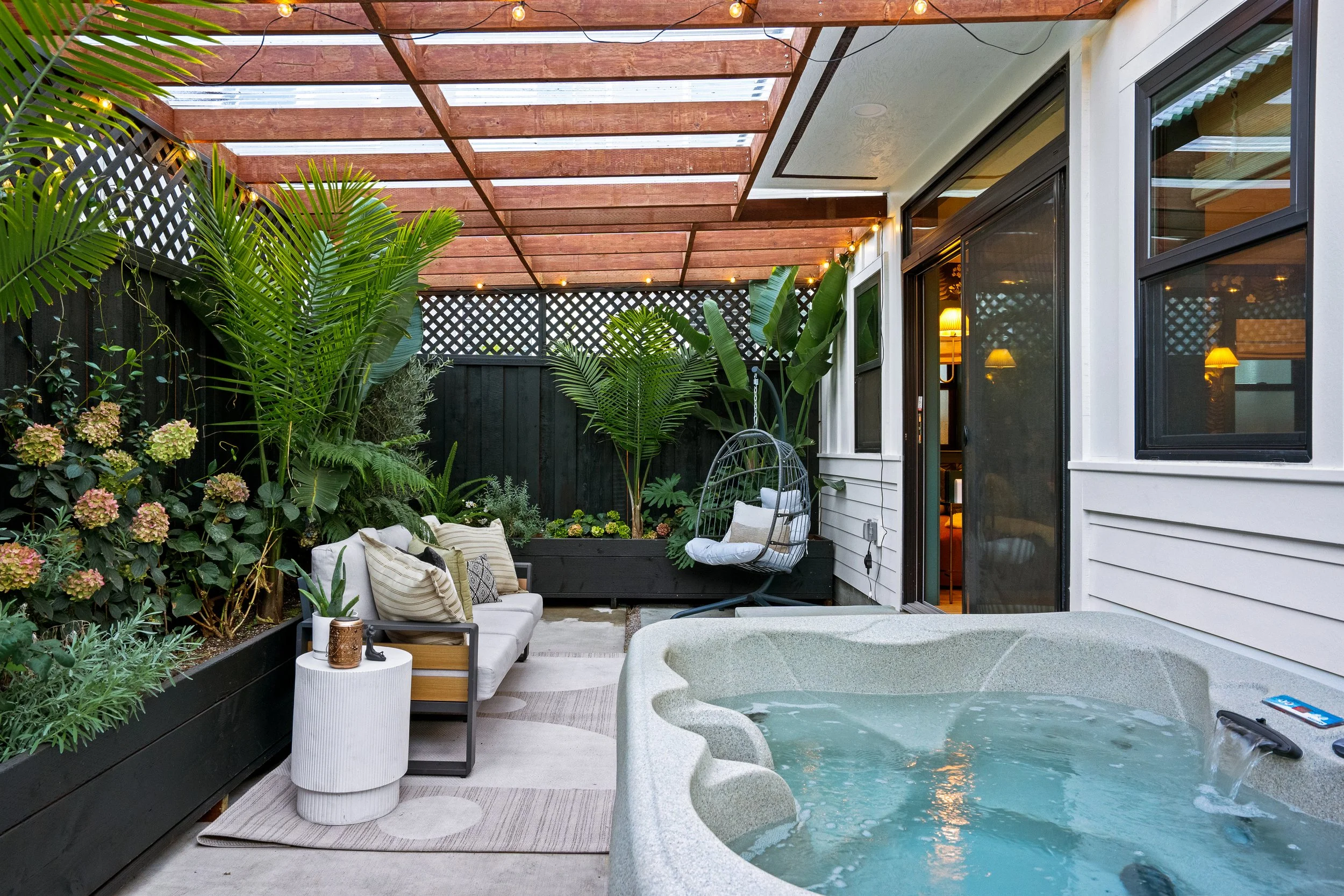 A cozy backyard patio with lush green plants, a beige sofa with pillows, a small white side table with decorative items, a black fence, string lights on a wooden pergola, a hanging egg chair, and a hot tub with water.