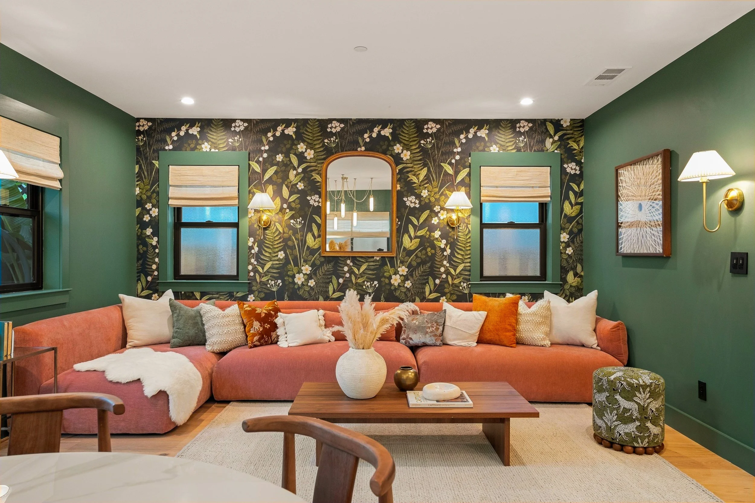 Living room with green walls and floral wallpaper, featuring a pink sectional sofa with assorted pillows, two wall-mounted lamps, a mirror, and a wooden coffee table with vases, in front of large windows with beige Roman shades.