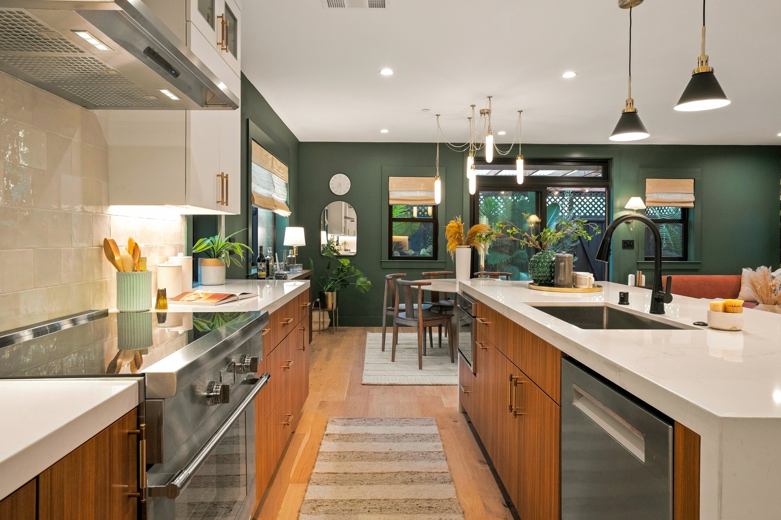 A modern kitchen with a green accent wall, wooden cabinets, white countertops, and black pendant lights. There are plants, a mirror, and a dining area with a table and chairs.