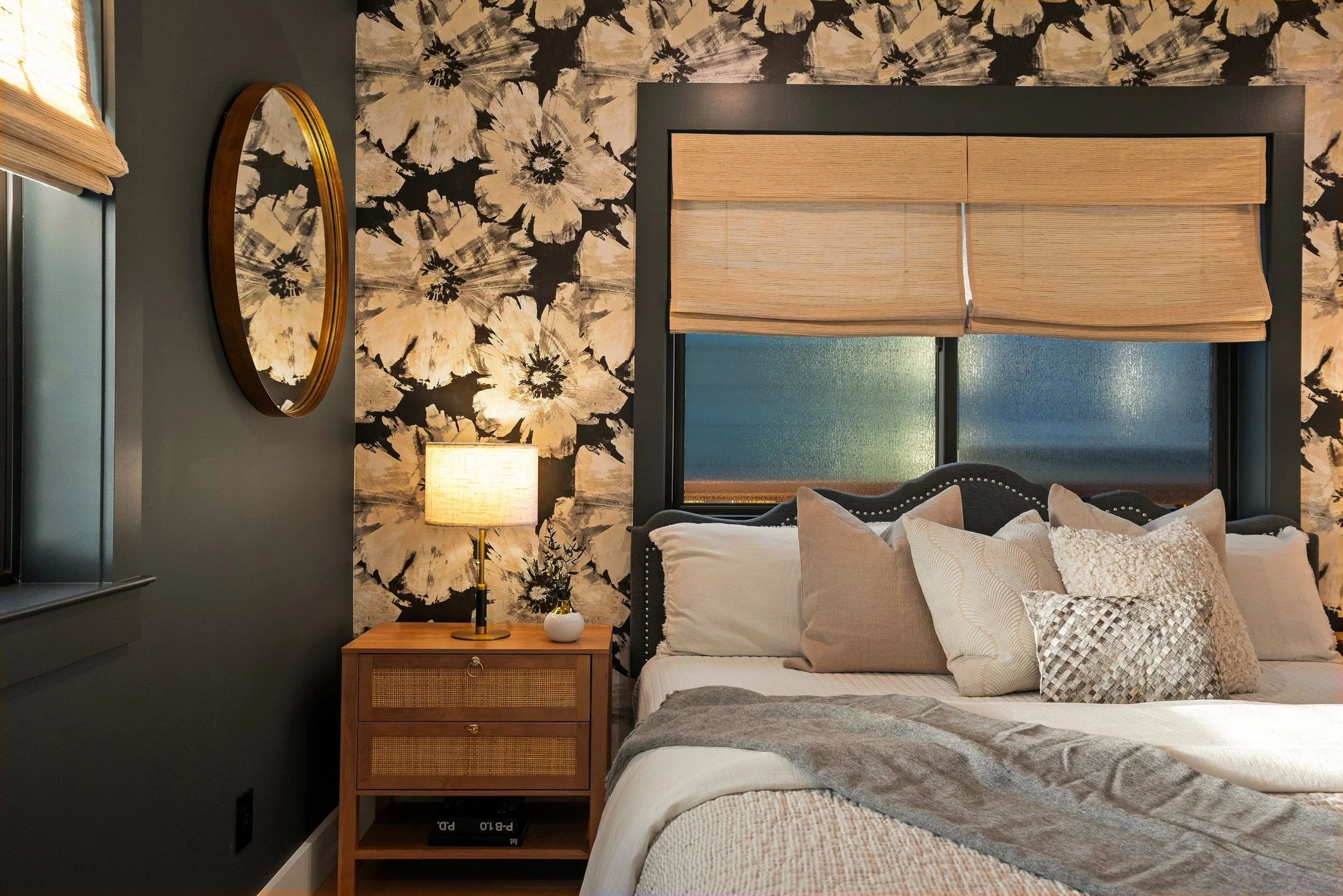 A cozy bedroom with a bed made with white and beige pillows and a gray throw blanket. There is a window with bamboo blinds, a dark gray wall, a wooden nightstand with a small drawer and rattan door, a table lamp, a small white vase with foliage, and 