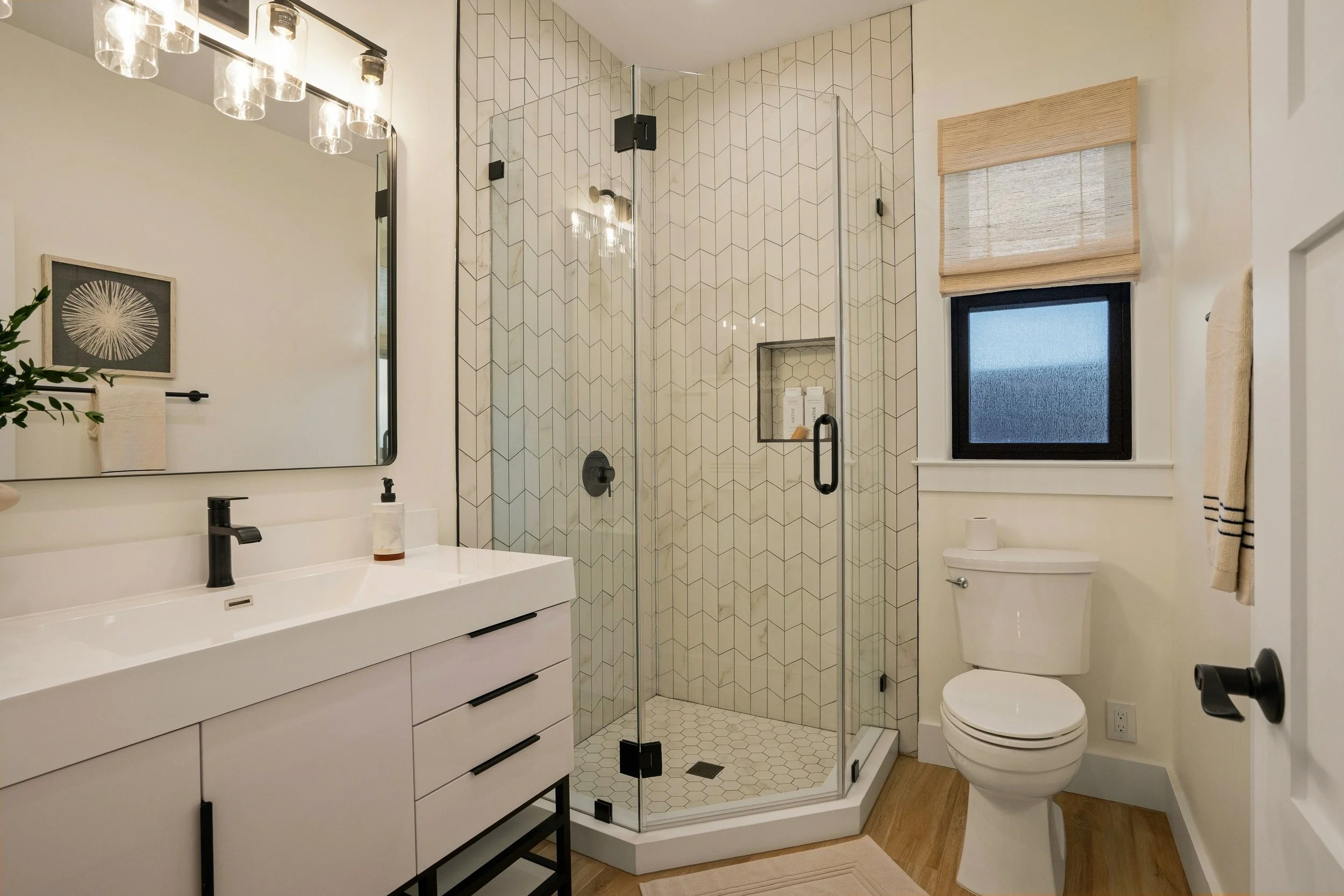 Bathroom with modern design, featuring a white vanity with black handles and a large mirror above, a glass-enclosed shower with geometric tile pattern, a small window with a bamboo shade, toilet, and various decor items like a framed artwork and a to