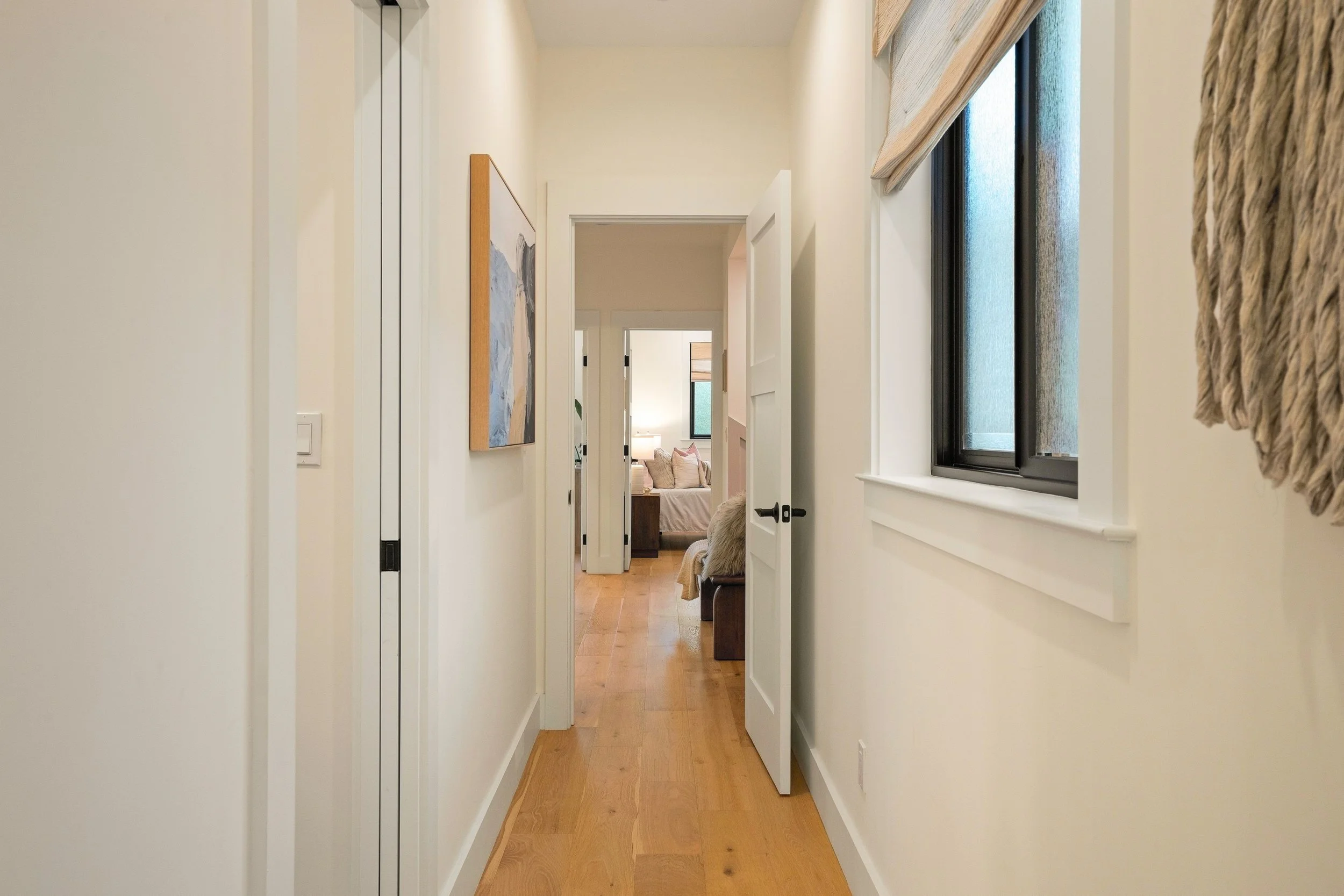 A hallway with cream-colored walls, wooden flooring, and white doors. Natural light coming through black-framed windows on the right, with a textured curtain on the window. Artwork on the left wall and a view into a living room with a sofa and a wind