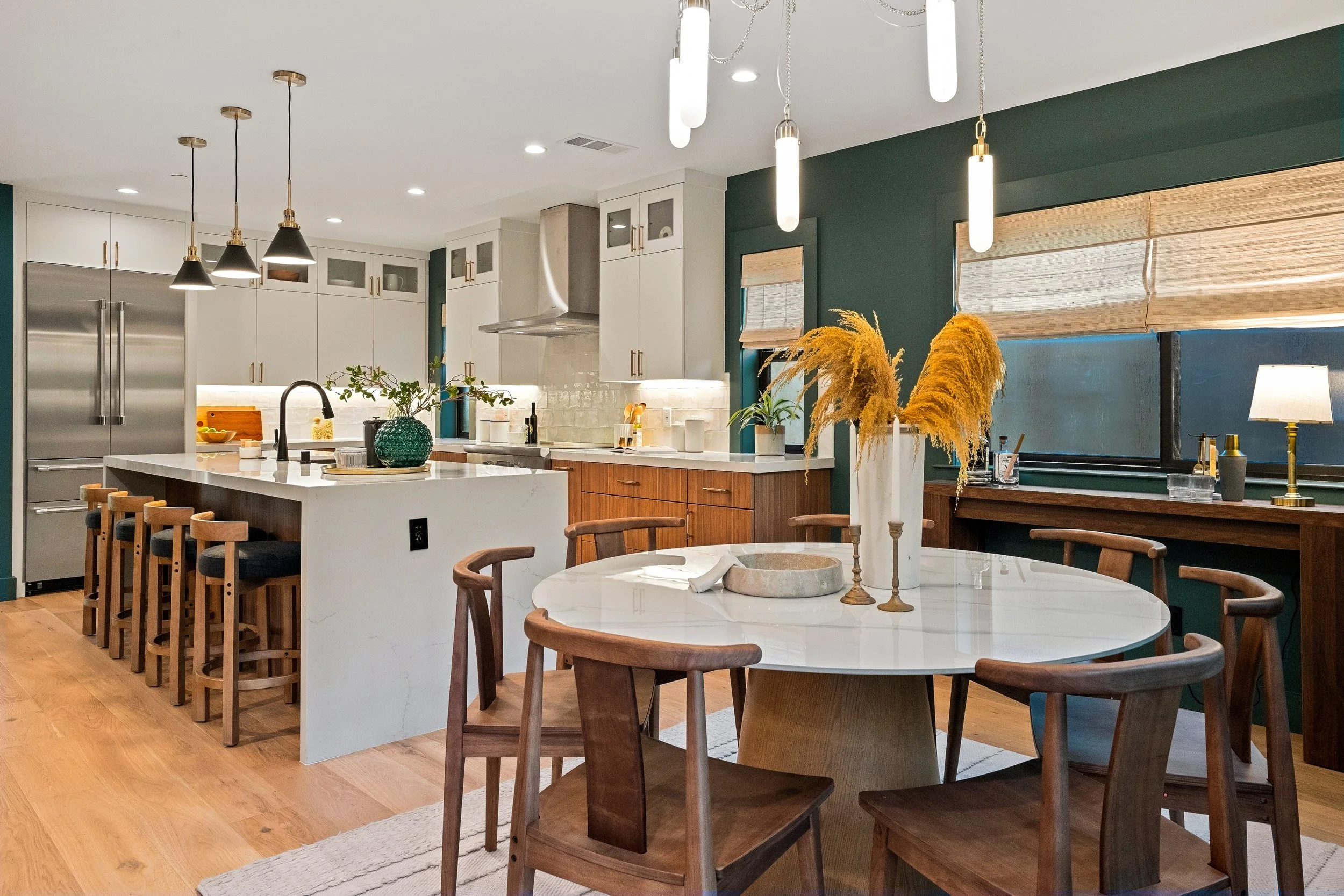 Modern kitchen with white cabinets, stainless steel refrigerator, wooden flooring, and a white island with bar stools. Decor includes a round dining table with wooden chairs, a vase with yellow pampas grass, and hanging and table lamps.