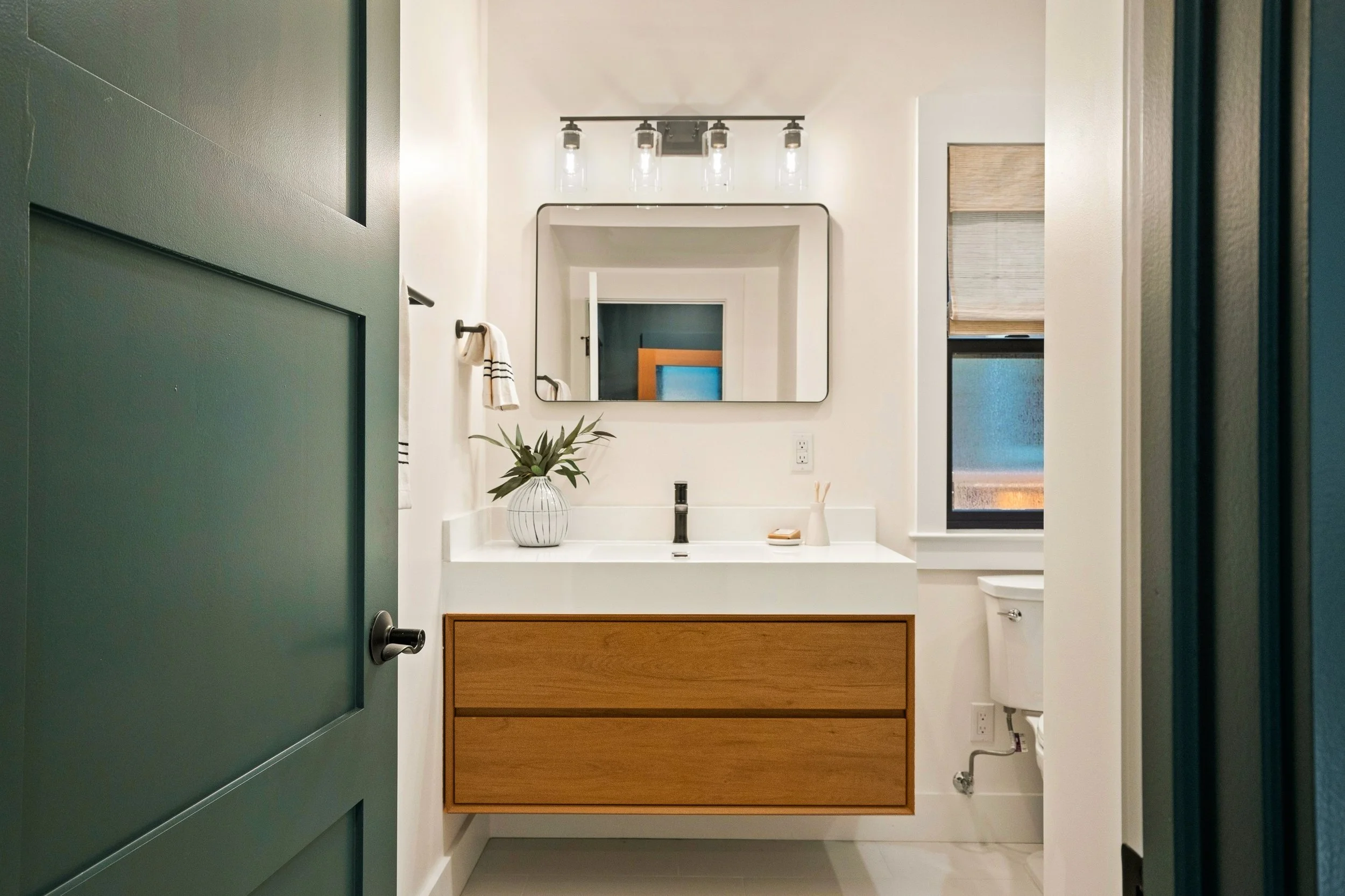 A view into a bathroom taken from a green door. Inside, there is a white vanity with a wooden drawer below. A potted plant is on the vanity. Above, a black-framed mirror and a modern light fixture with five bulbs are visible. On the left wall, a towe