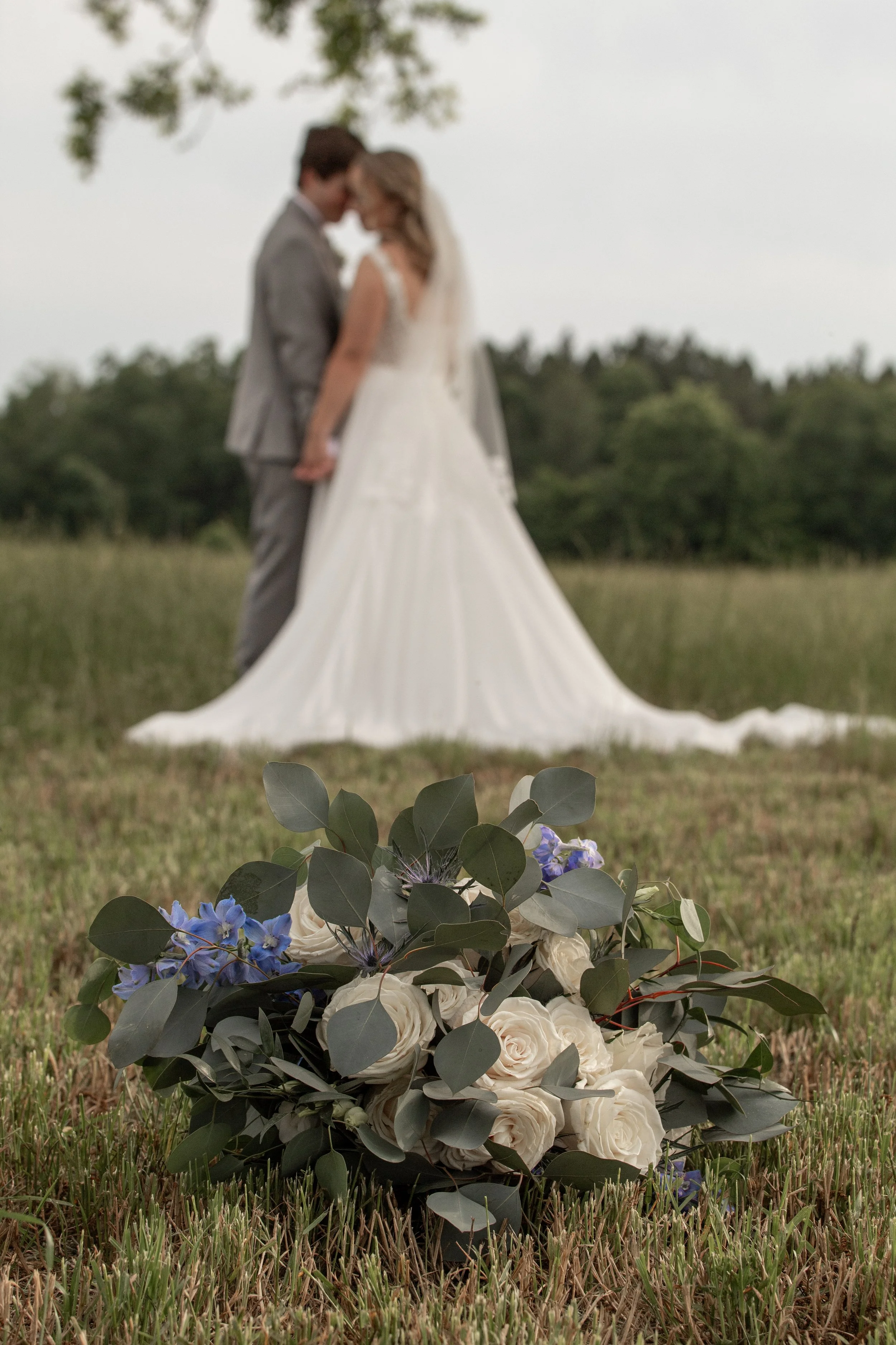 Couple kissing outdoors at their wedding, with the bride in a white gown, groom in a gray suit, and a floral arrangement in the foreground.
