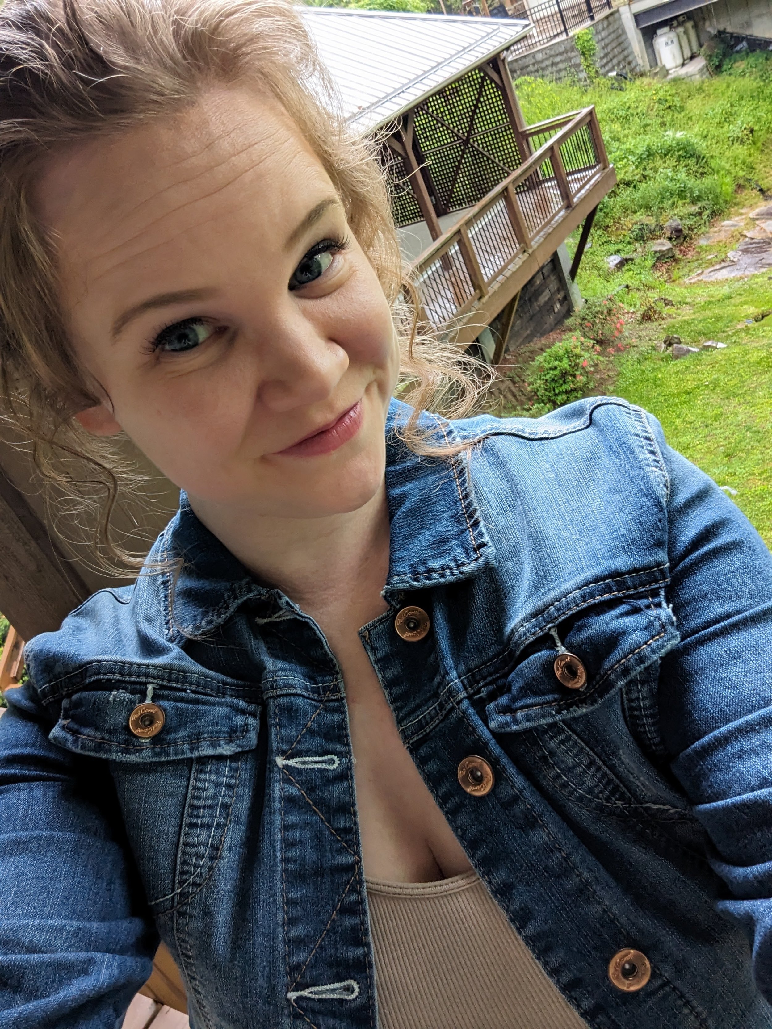 Close-up selfie of a smiling blonde woman in a denim jacket outdoors near a wooden deck and landscaped garden.
