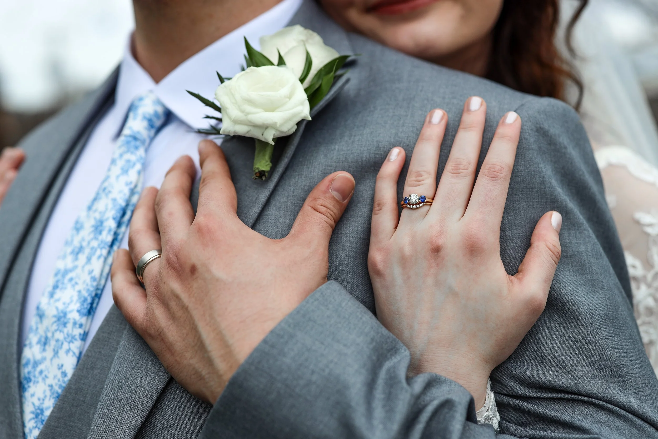 Close-up of a newlywed couple embracing, with white roses, wedding rings, and a sapphire engagement ring visible.