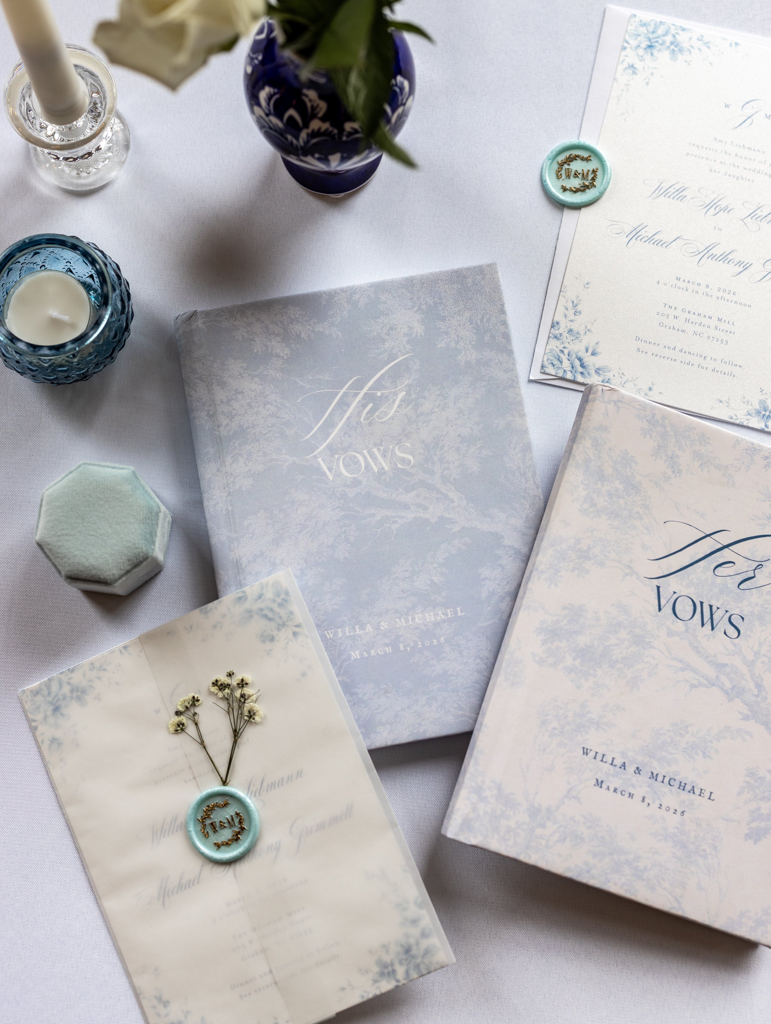 Flat lay of elegant wedding stationery, floral invitations, wax seals, candles, and blue-and-white floral decor on a white tablecloth