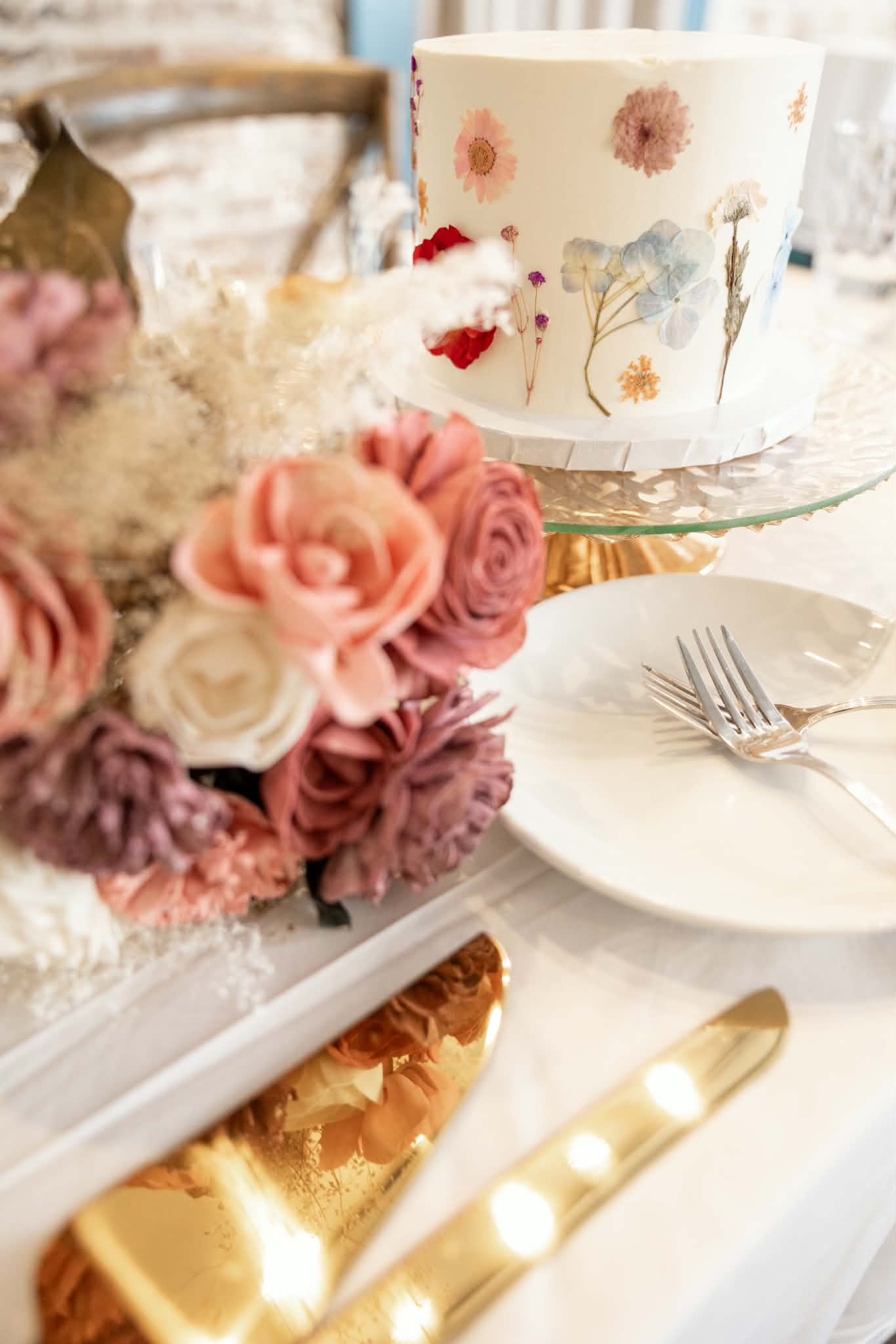 Elegant table setting with a floral-decorated cake, pastel rose centerpiece, white plates, and gold flatware