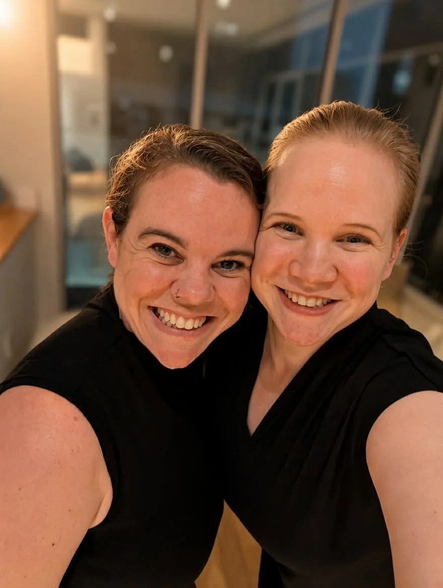 Two smiling women in black tops take a close-up selfie indoors.