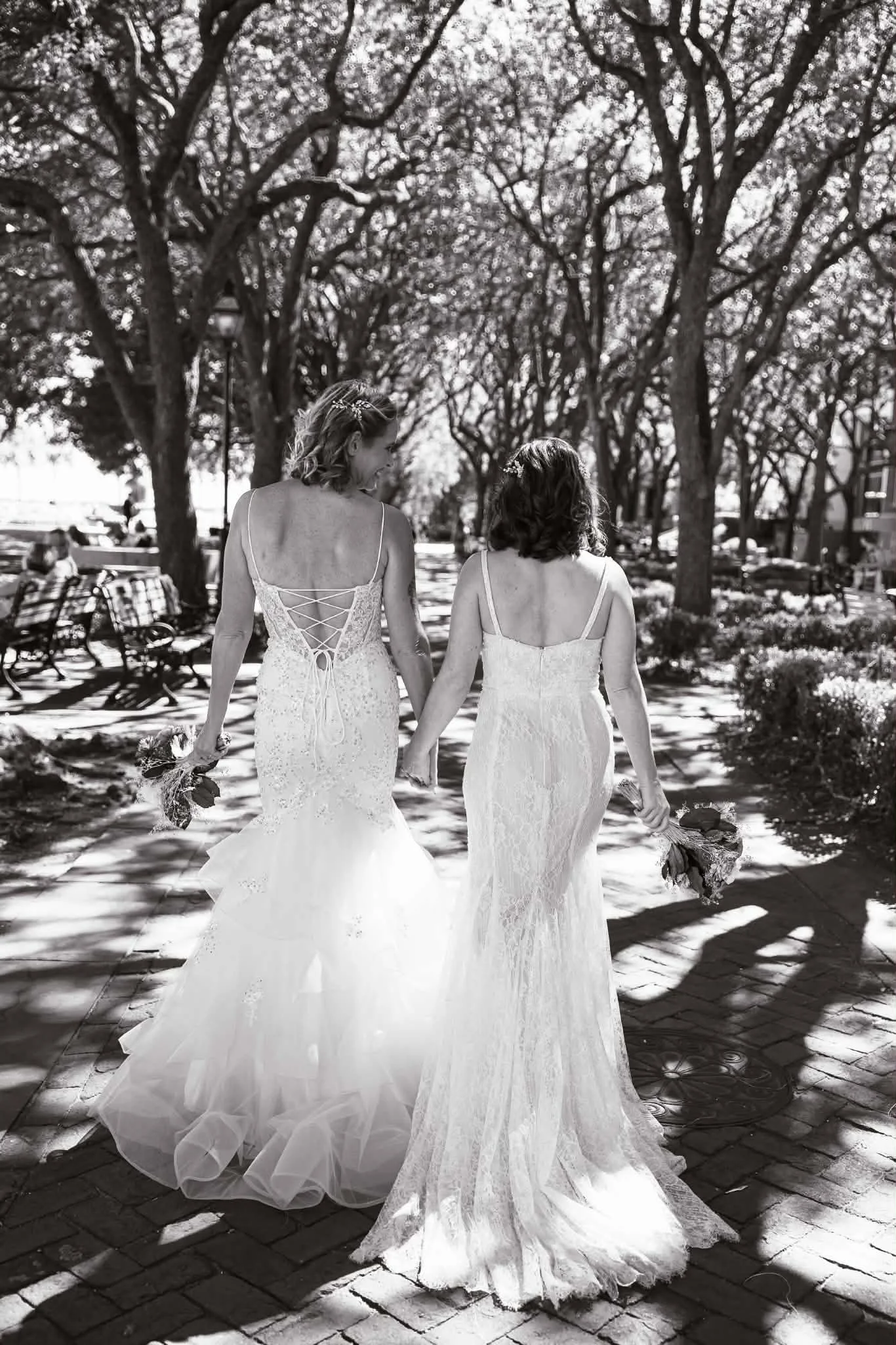 Black-and-white wedding photo of two brides in lace gowns walking hand in hand through a tree-lined garden, carrying bouquets.
