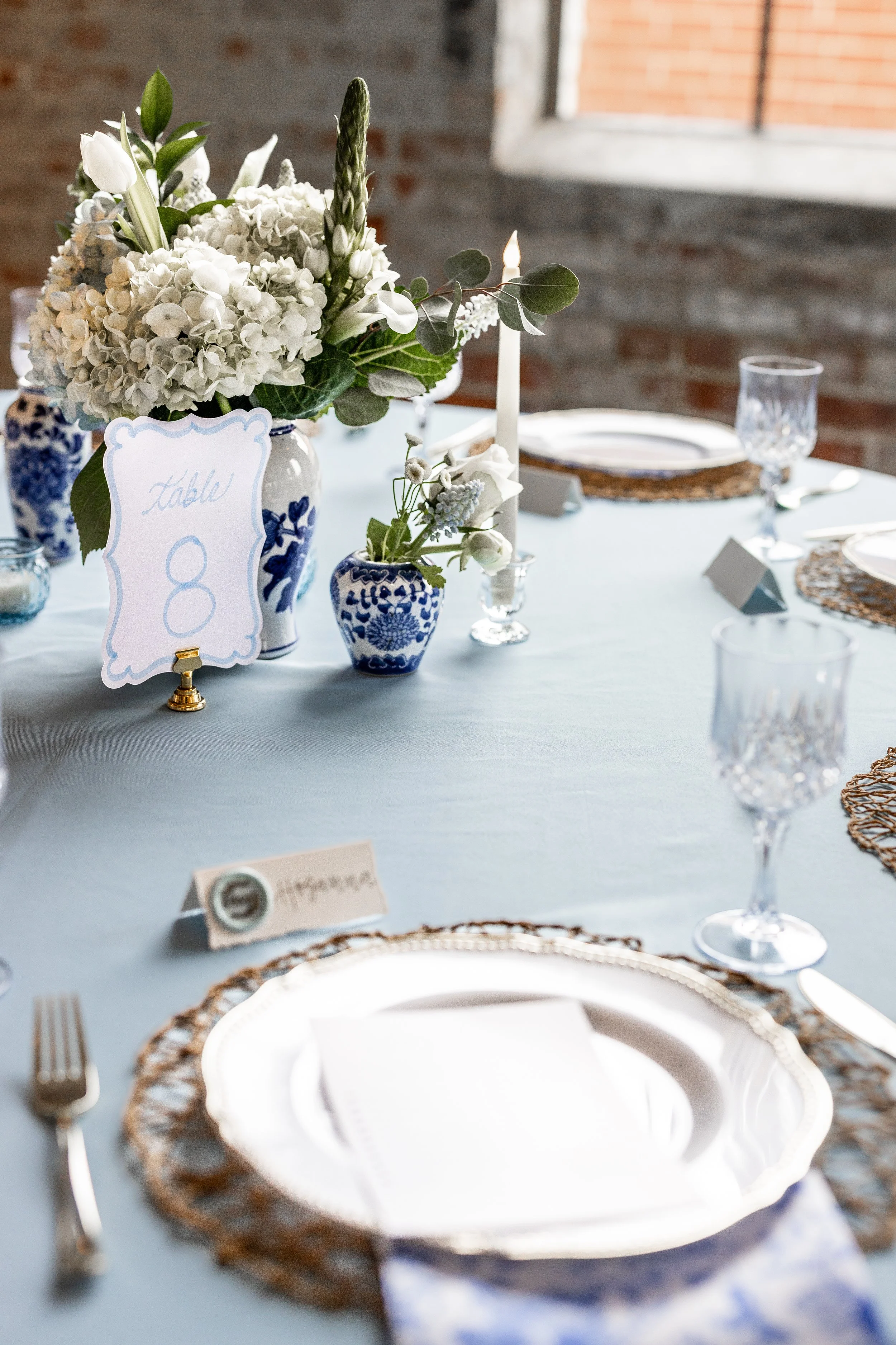 Elegant wedding table setting with white floral centerpiece, blue-and-white vases, lit candles, place cards, china plates, and glassware