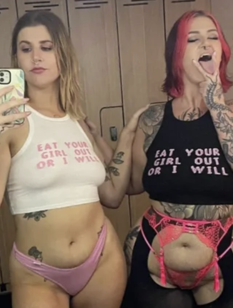 Two women taking a mirror selfie in a room with wooden cabinets. The woman on the left is wearing a white crop top and pink underwear. The woman on the right has pink and black hair, tattoos, and is wearing a black crop top with pink text, and pink lace underwear with garters.