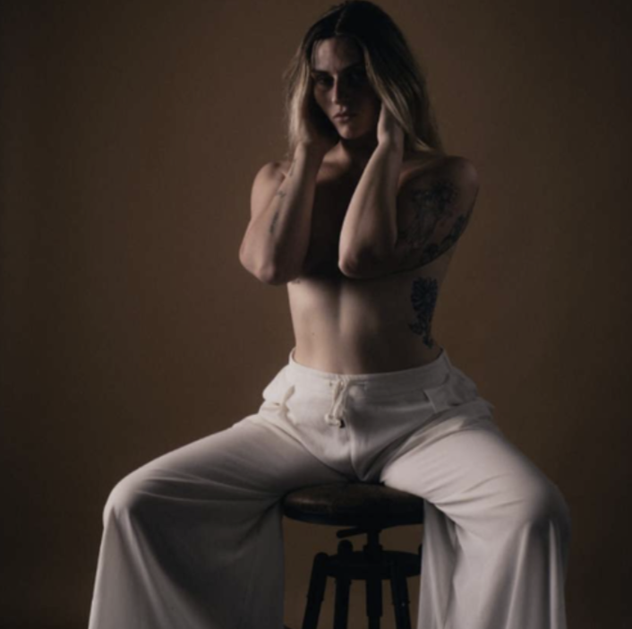 A topless woman with tattoos sits on a stool against a dark background, wearing wide-leg white pants and posing with her hands near her face.