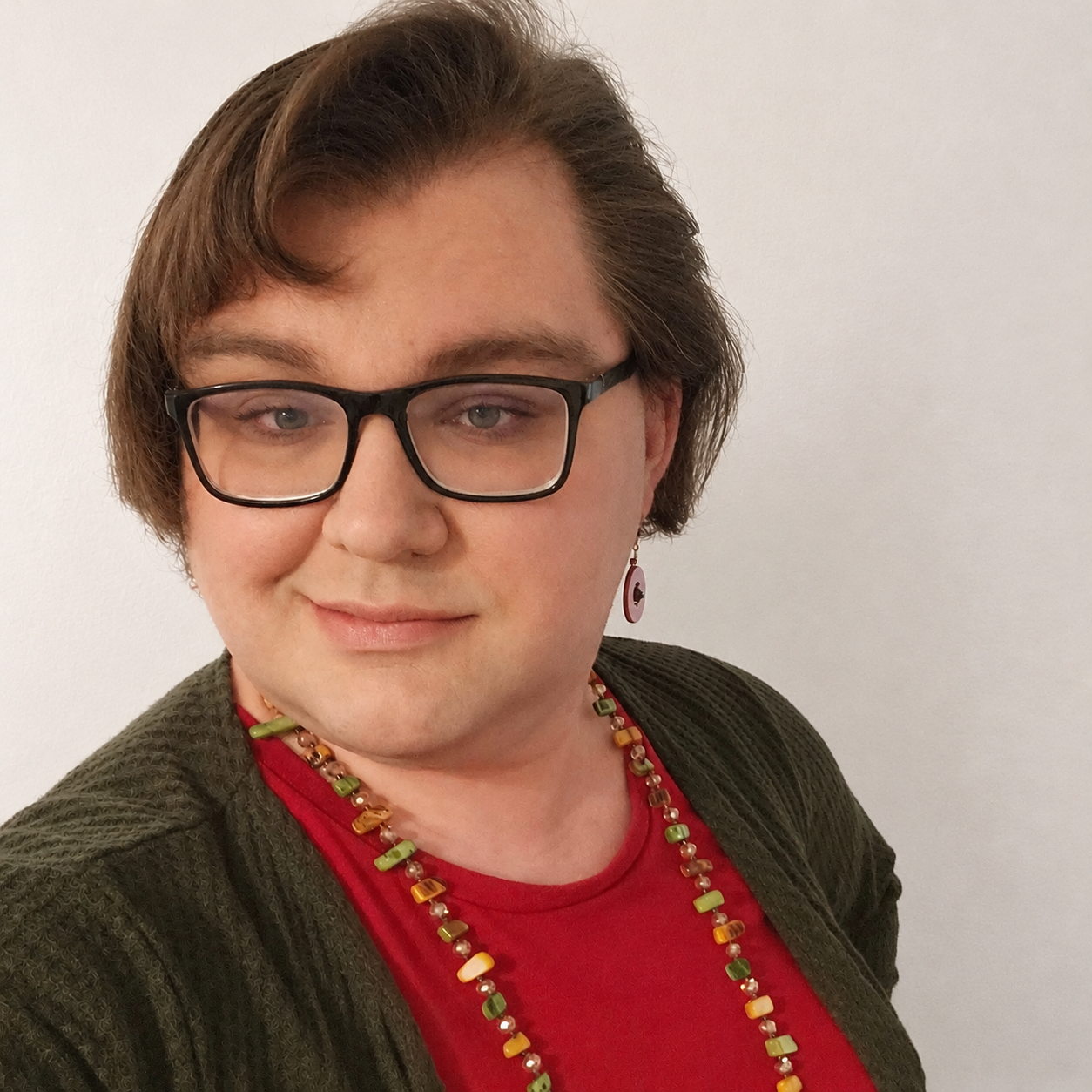 A person with short brown hair and glasses, wearing a red shirt, green cardigan, colorful beaded necklace, and earrings, smiling at the camera against a plain white wall.